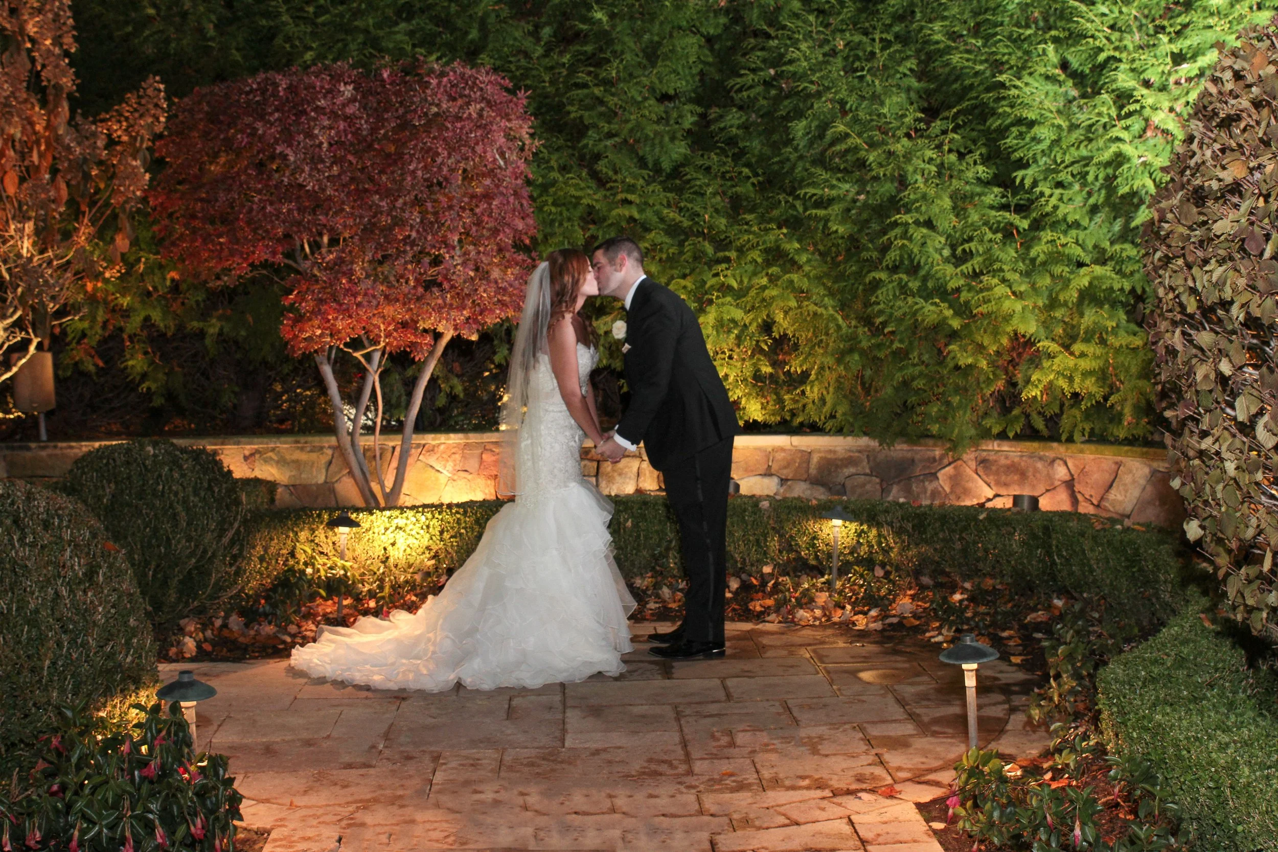 A bride and groom kissing at night outdoors in a garden, with trees and shrubs illuminated by small landscape lights.