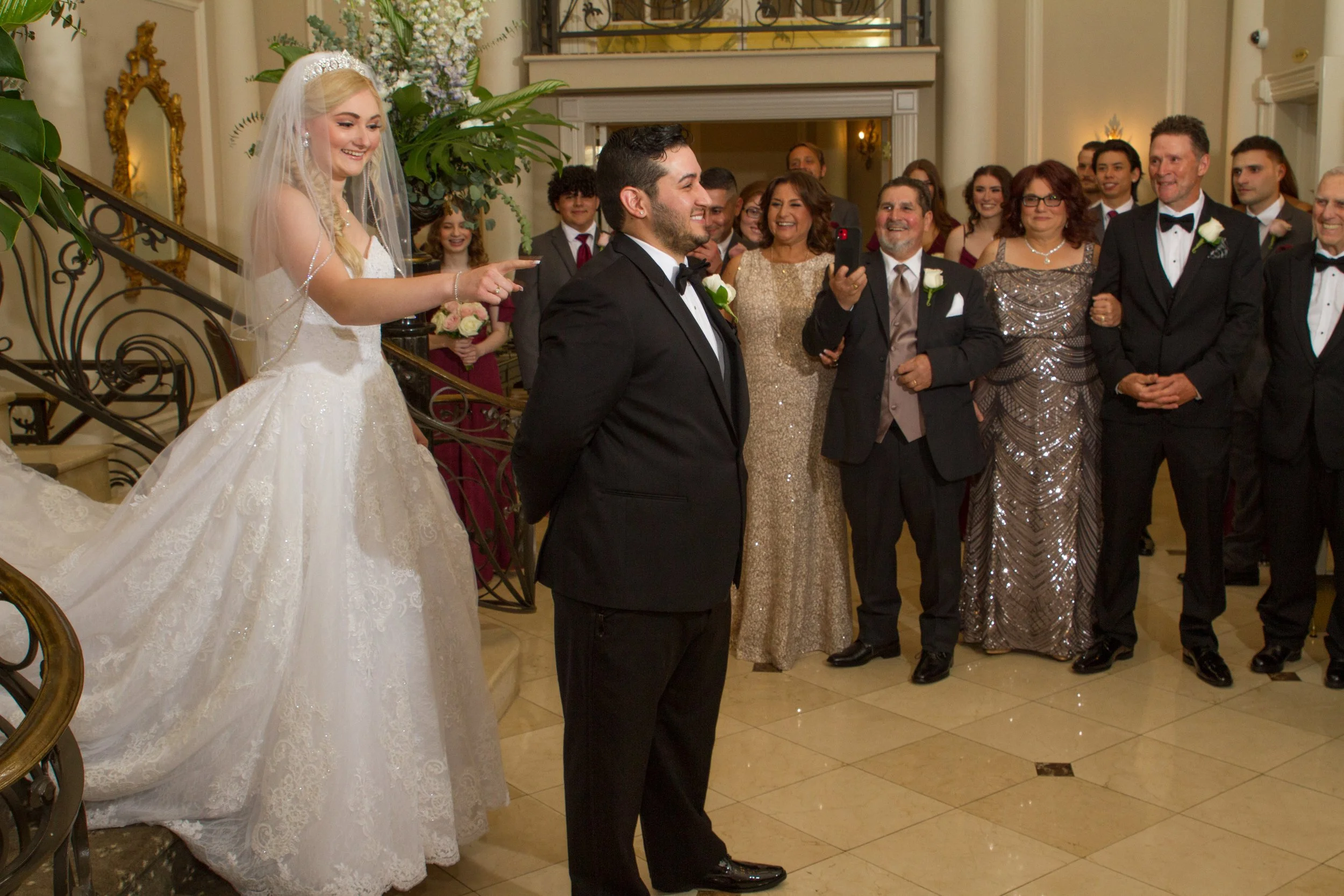 A bride and groom are standing in the center of a wedding reception, with the bride pointing and the groom smiling. Guests in formal attire are gathered around, smiling and taking photos inside an elegant venue with floral decorations.