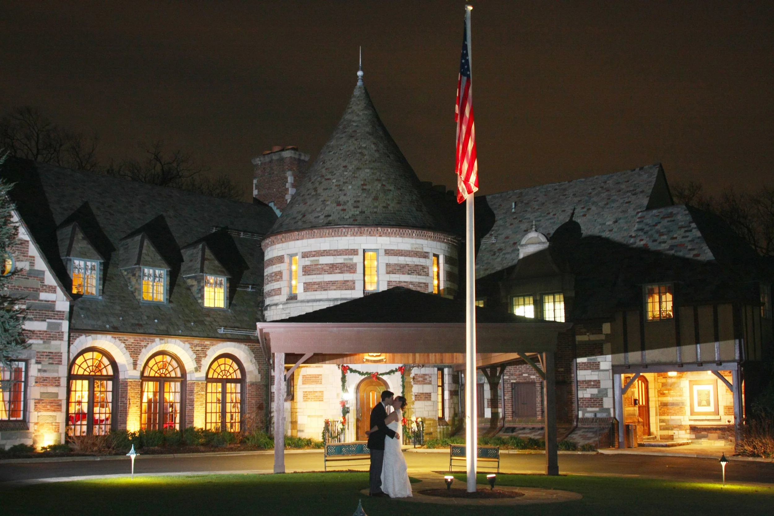 A couple in wedding attire standing under a flagpole with an American flag in front of a large, historic stone and brick building at night, decorated with holiday wreaths and lighting at the Ridgewood Country Club.