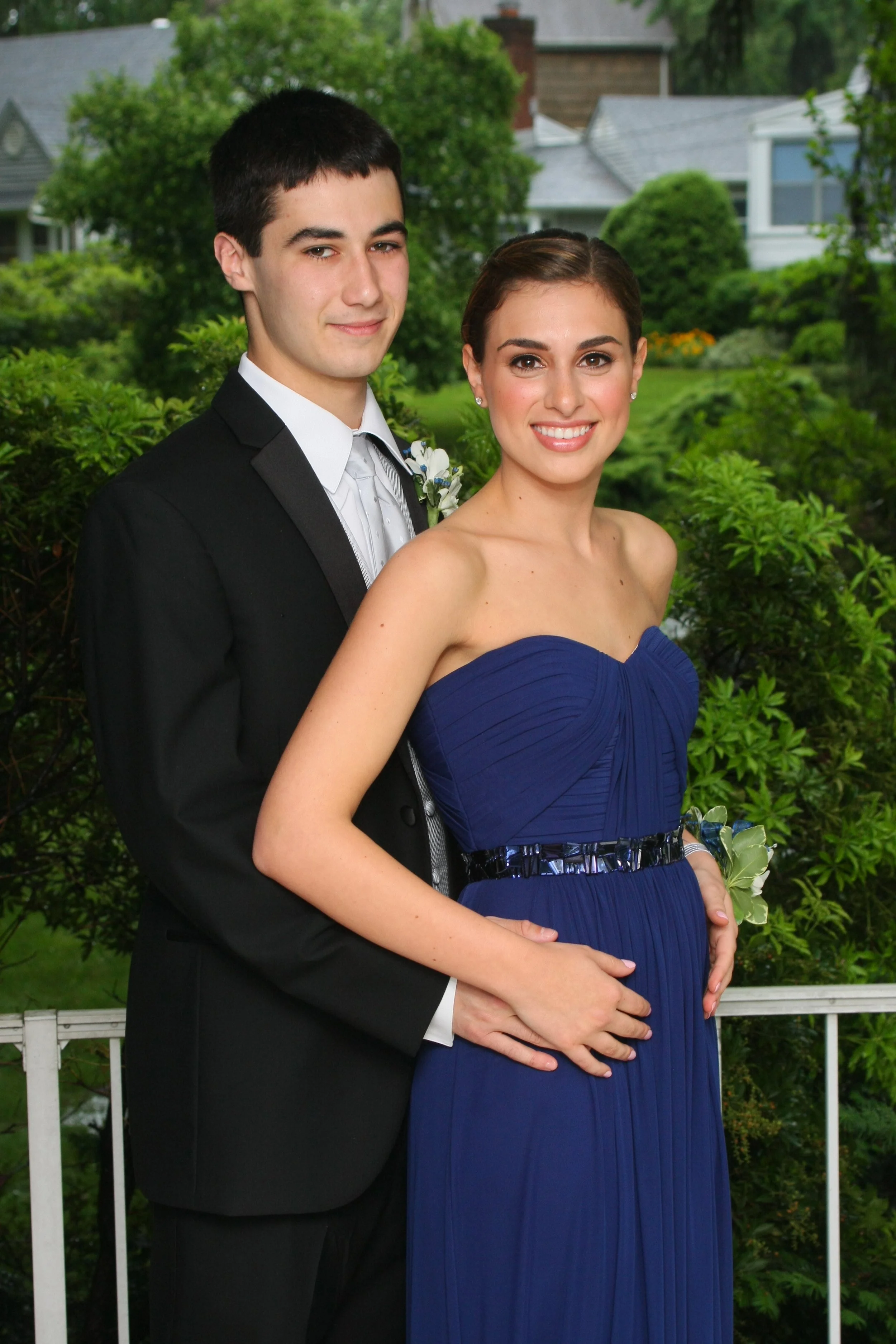 A young couple posing outdoors on a balcony with a green garden and houses in the background. The man is dressed in a black tuxedo with a white shirt and tie, and the woman is wearing a strapless, royal blue gown. The woman is holding her pregnant be