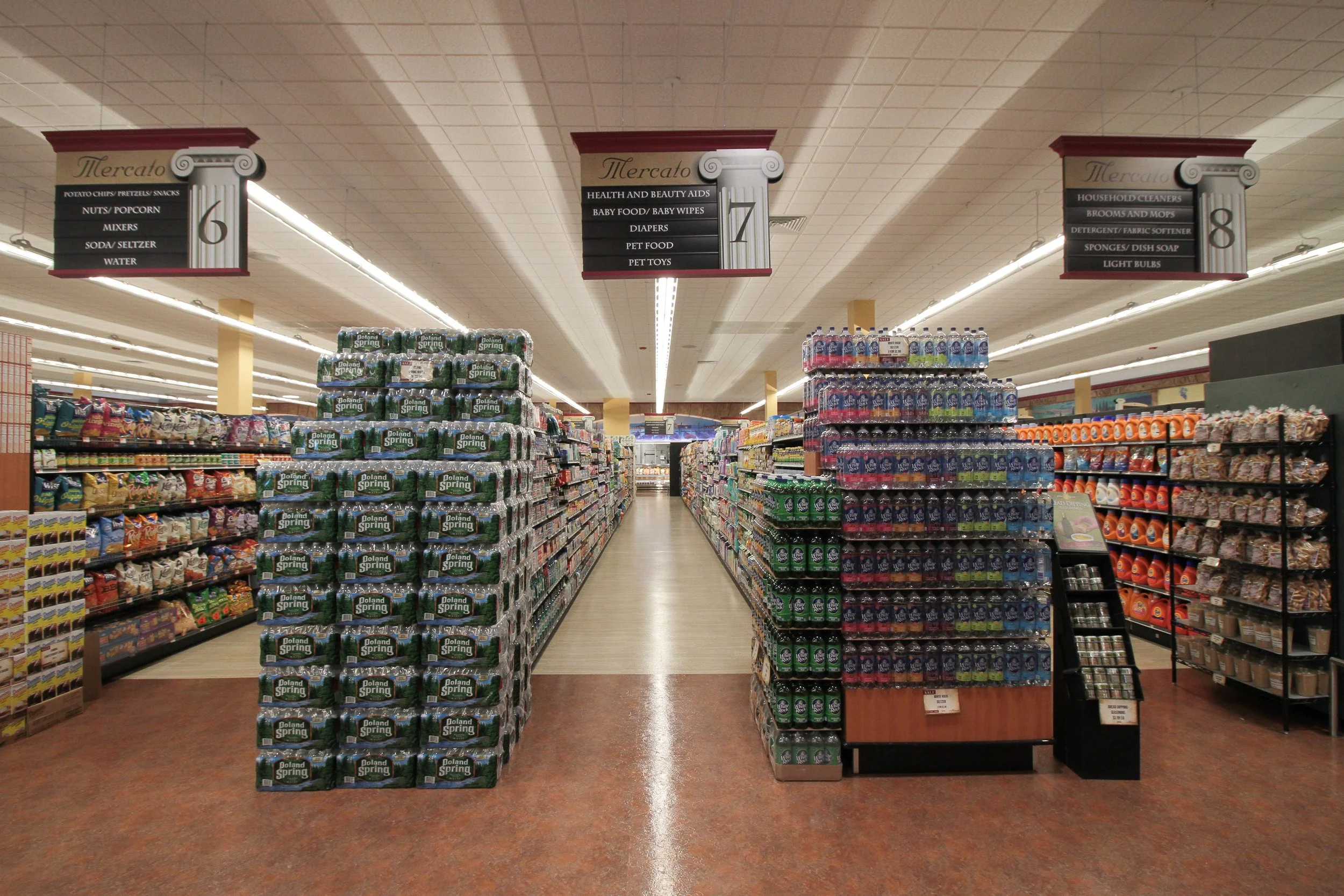 Aisle in a grocery store with shelves stocked with bottled water, snacks, and household items. Overhead signs indicating different sections such as groceries, health and beauty aids, and household cleaners.