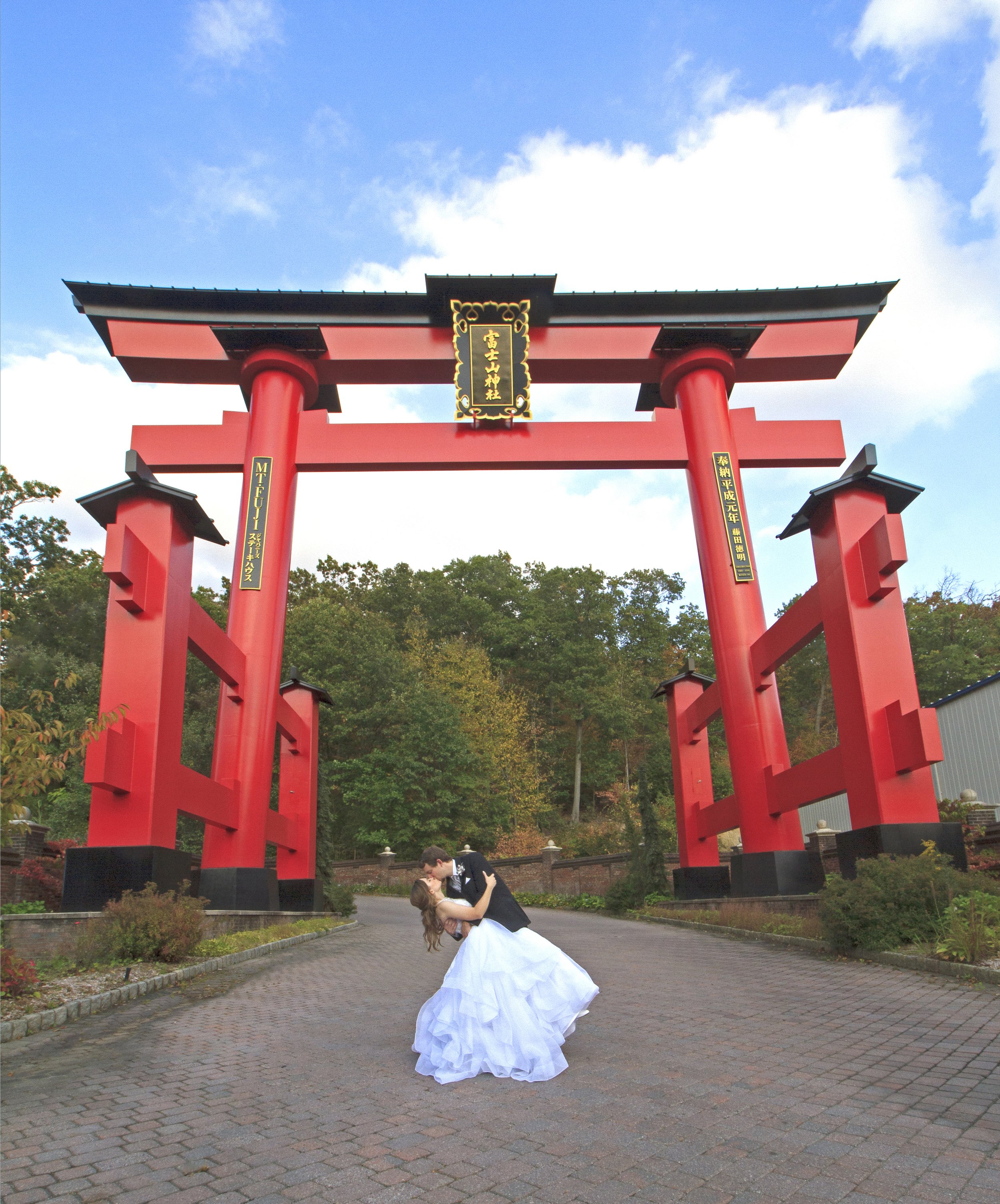 A bride and groom share a kiss beneath a large, red traditional Japanese torii gate outdoors at the Views at Mount Fuji on a paved path, with trees and a cloudy sky in the background.