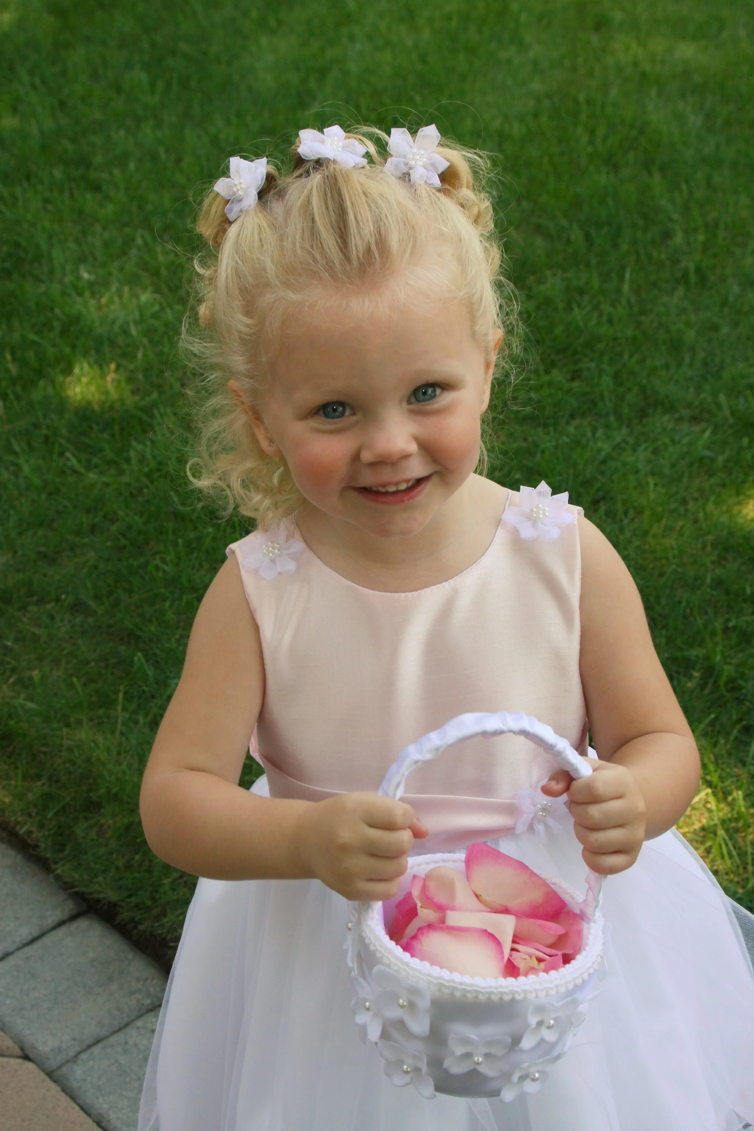 A young girl with blonde curly hair, dressed in a light pink and white dress, holding a white basket filled with pink flower petals, standing on a sidewalk next to a grass lawn.
