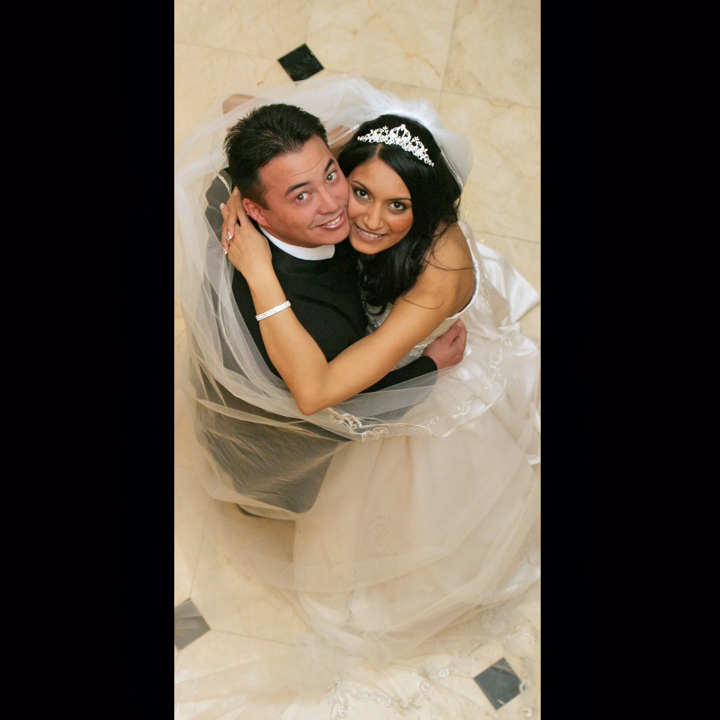 A bride and groom embracing and smiling at the camera, taken from above. The bride wears a white wedding dress and tiara, and the groom wears a black tuxedo with a white collar, on a tiled floor.