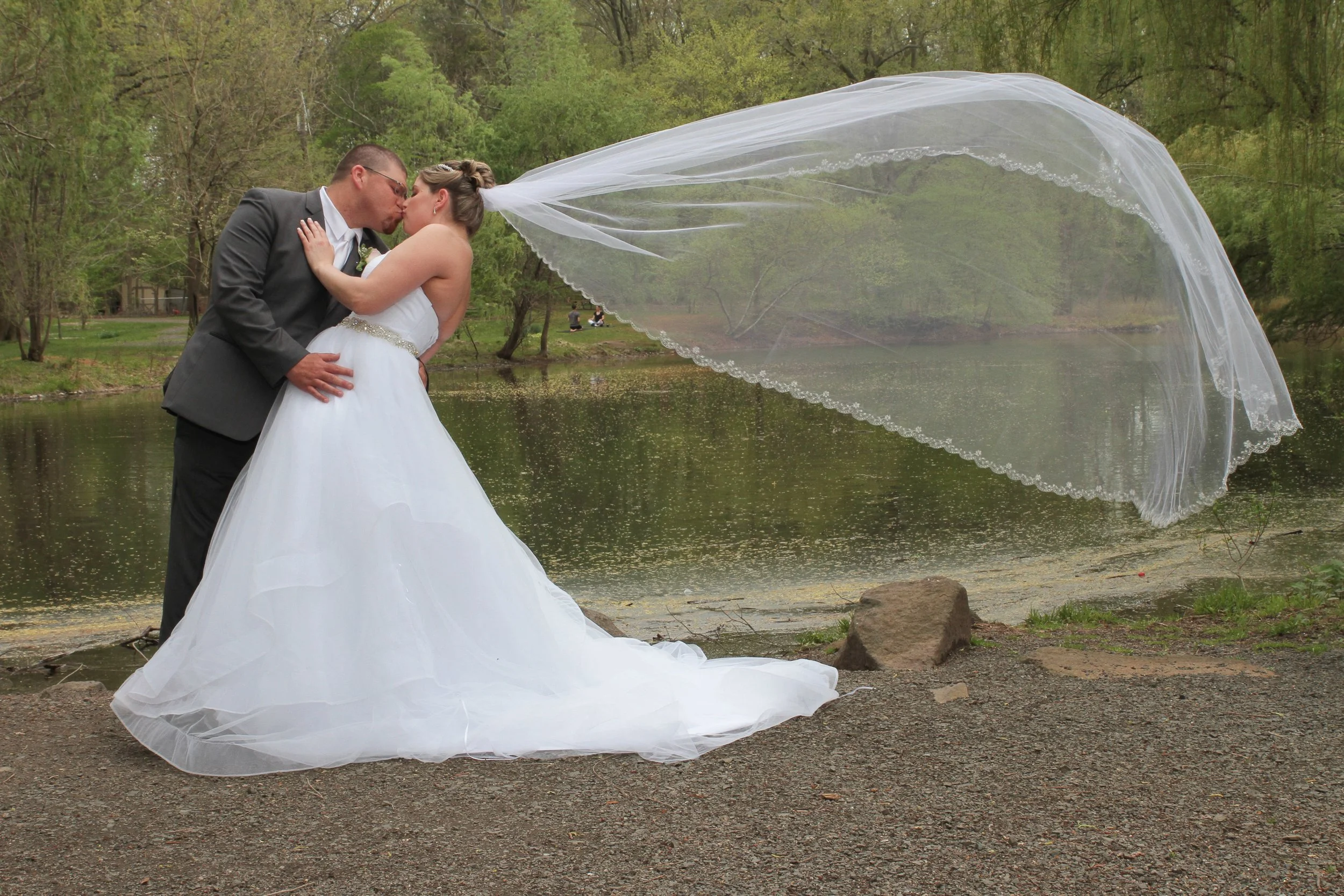 A bride and groom kissing by a lake with greenery in the background, the bride's veil flowing in the wind.