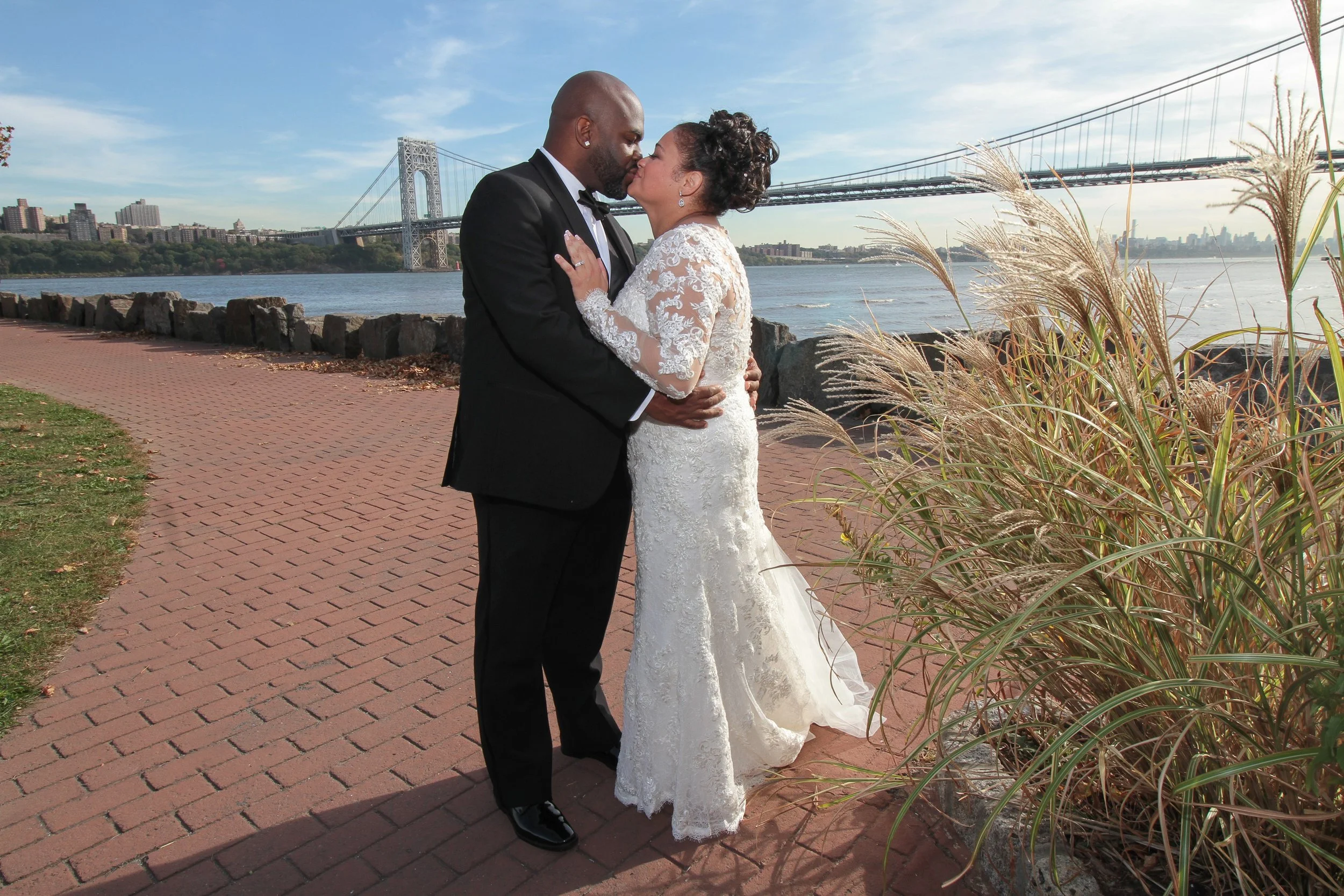 A couple dressed in wedding attire sharing a kiss by the water with a city skyline and bridge in the background.