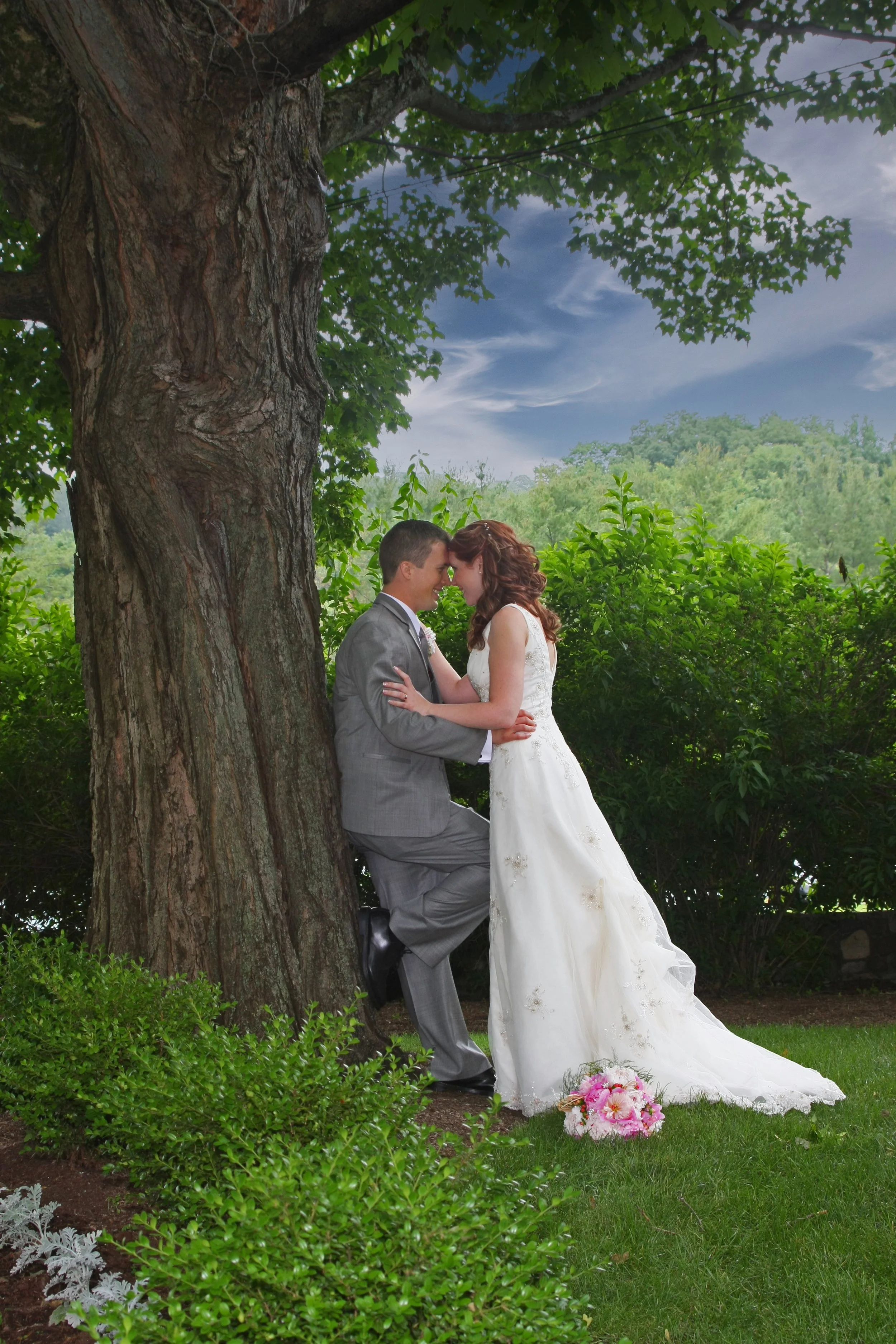 A bride and groom leaning against a large tree outdoors, touching foreheads, with lush green foliage and cloudy sky in the background. The bride is in a white wedding gown with a pink bouquet on the grass.