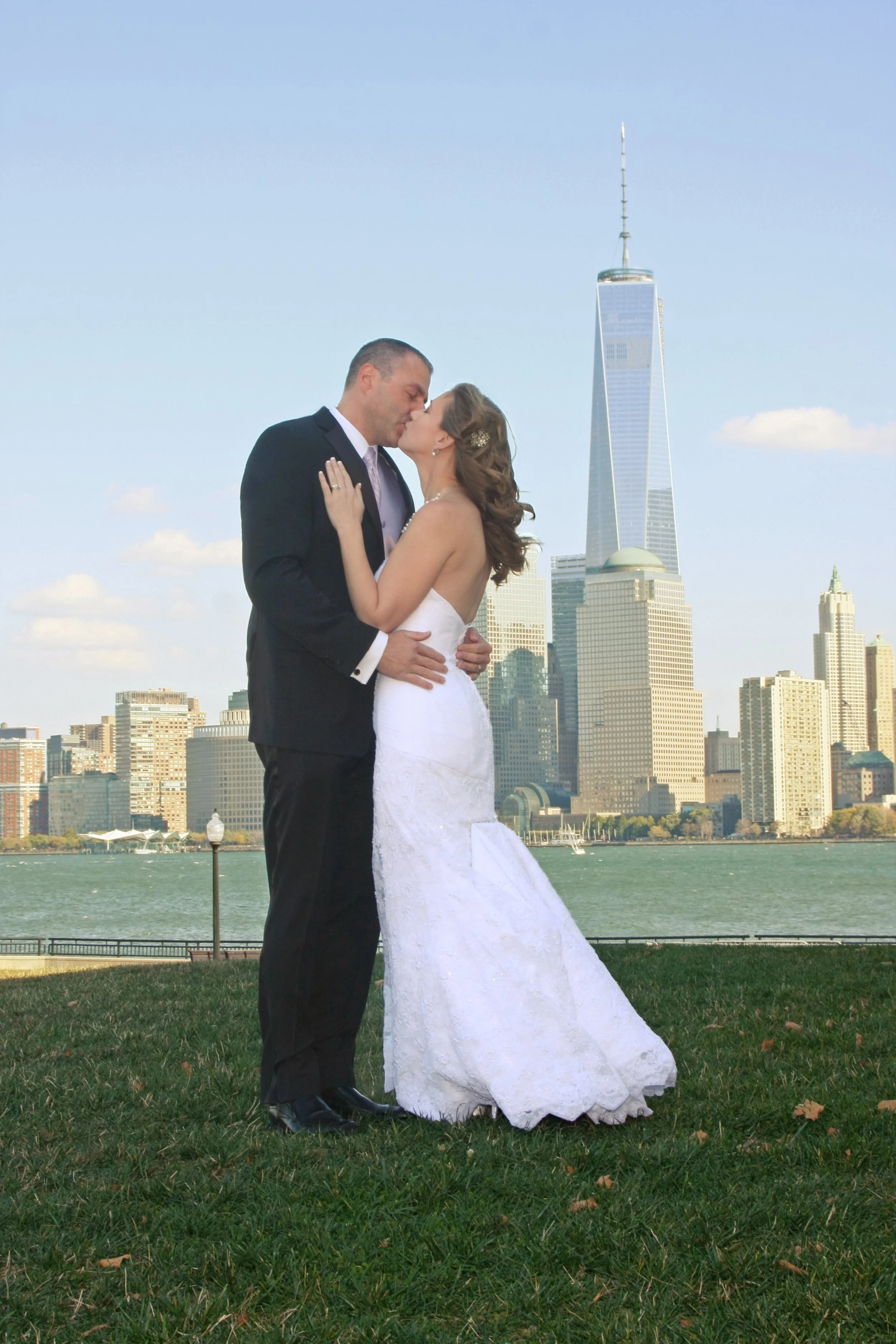 A bride and groom sharing a kiss outdoors with the New York City skyline, including the One World Trade Center, in the background.