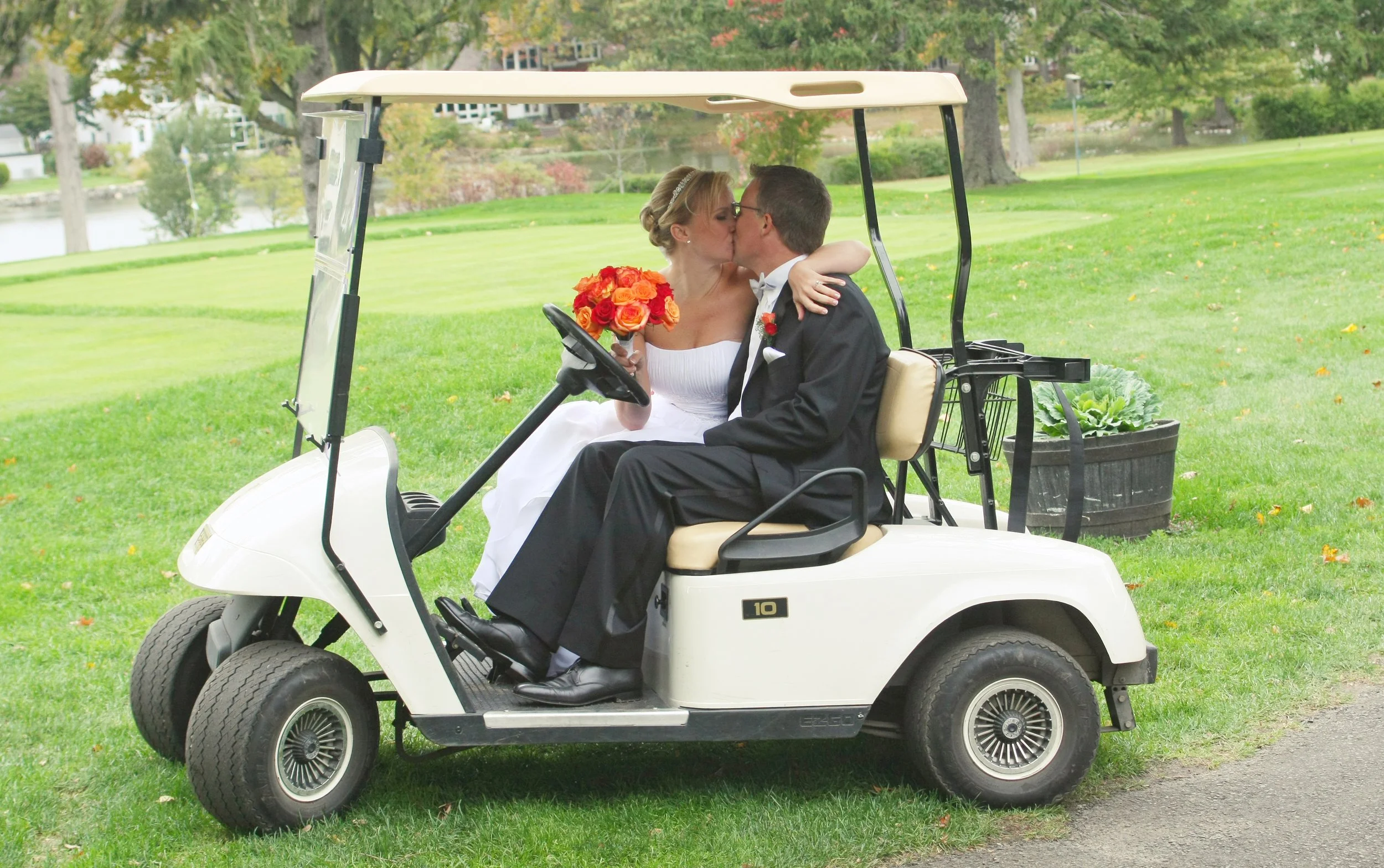 A bride and groom in wedding attire sitting in a golf cart, sharing a kiss. The bride holds a bouquet of orange and red flowers, and the background features a green lawn, trees, and water.