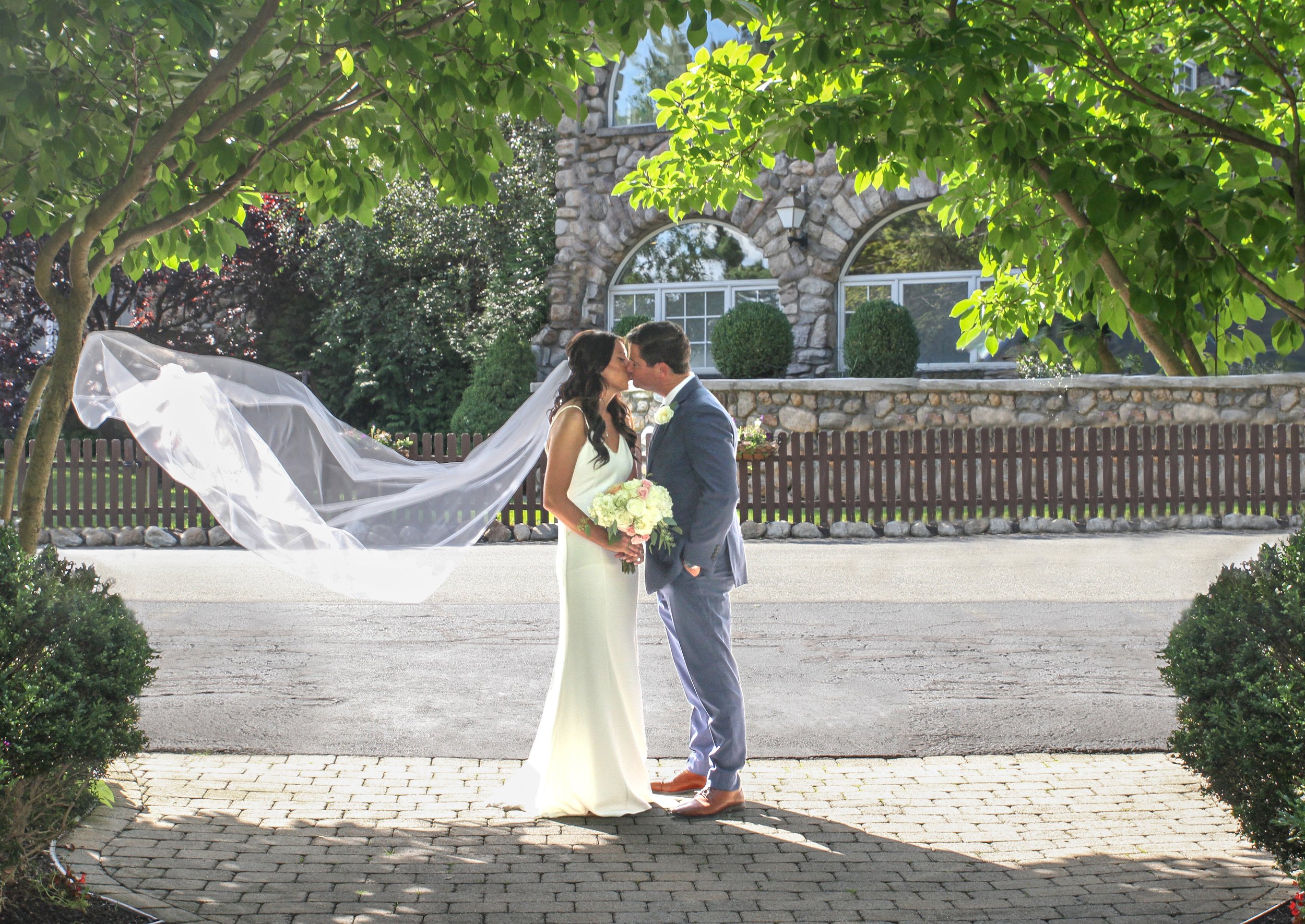 A bride and groom sharing a kiss outdoors during their wedding, with the bride holding a bouquet and her veil flowing behind her, framed by trees and a rustic house in the background at the Ramsey Golf and Country Club.