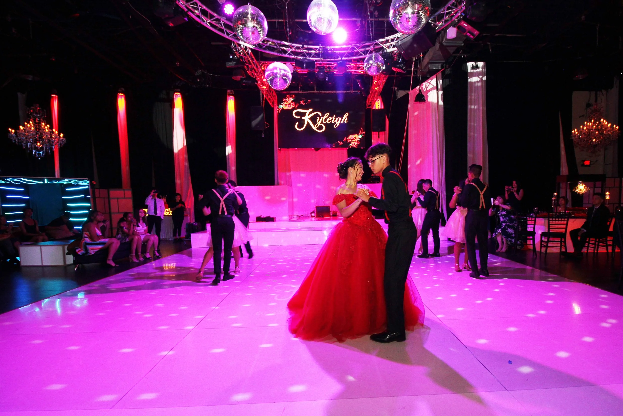 A sweet sixteen couple dances on a pink-lit dance floor in a ballroom decorated with chandeliers and disco balls, with guests watching and dancing in the background.