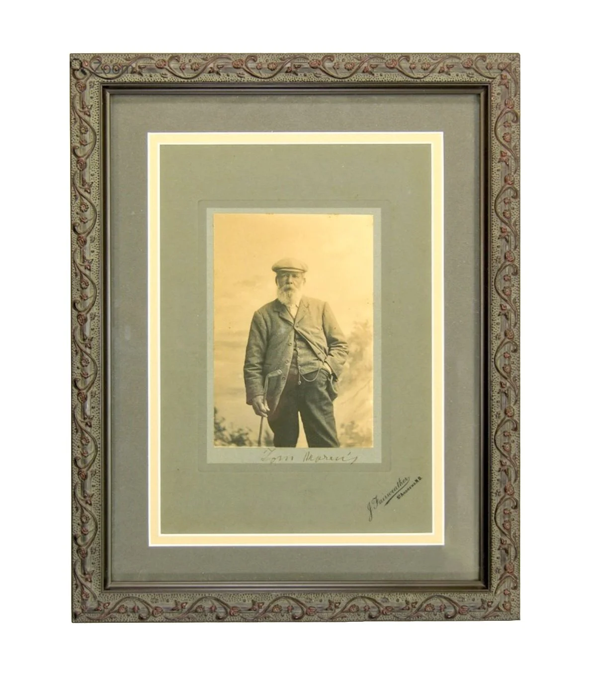 A framed black-and-white photograph of an older man with a beard, wearing a hat and vintage clothing, holding a cane, with trees in the background. The frame has ornate detailing and there are signatures or inscriptions on the mat around the photogra