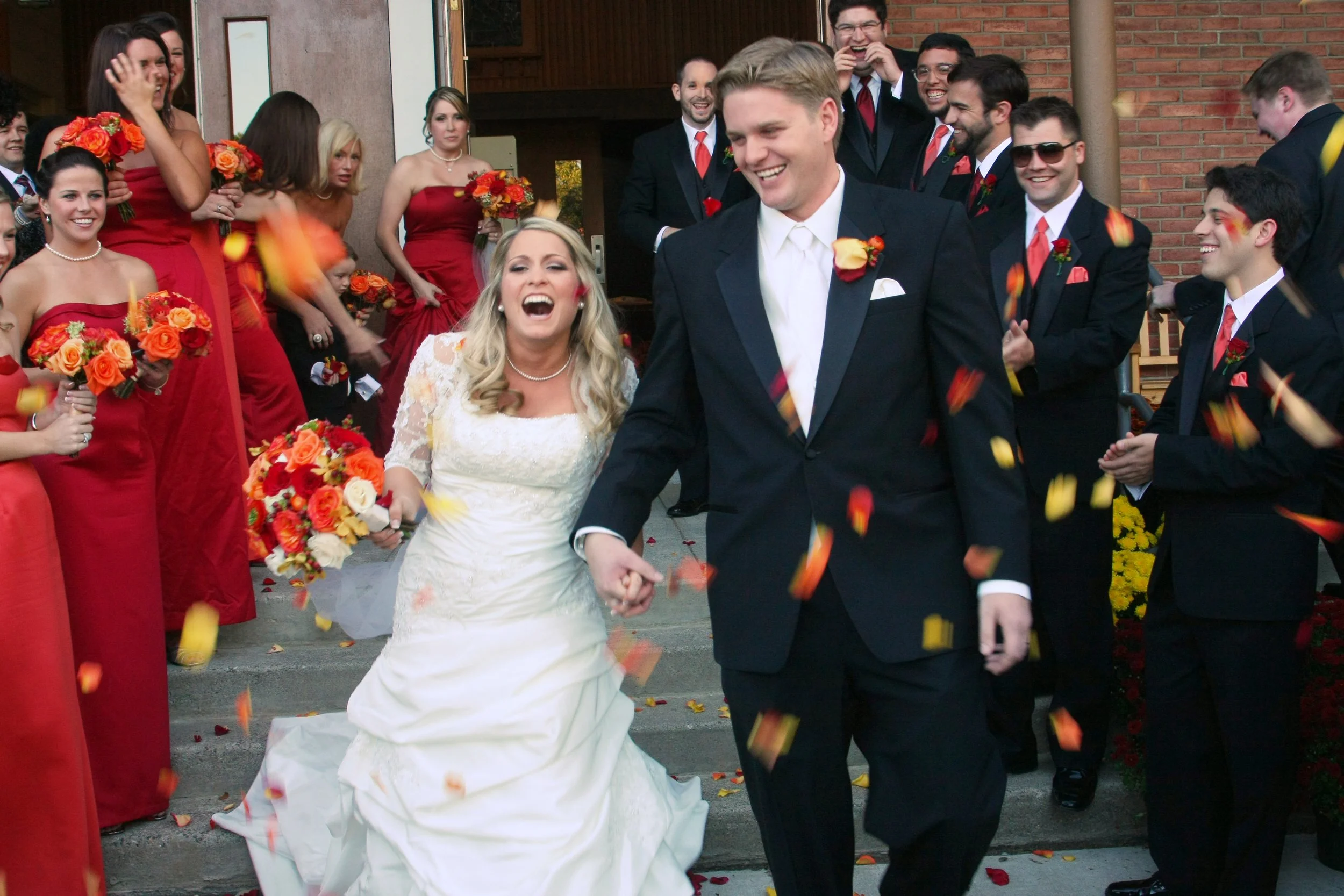 A newlywed couple is smiling and holding hands as they walk away from their wedding ceremony surrounded by friends and family. The bride is wearing a white wedding dress and holding a bouquet of red, orange, and white flowers. The groom is wearing a
