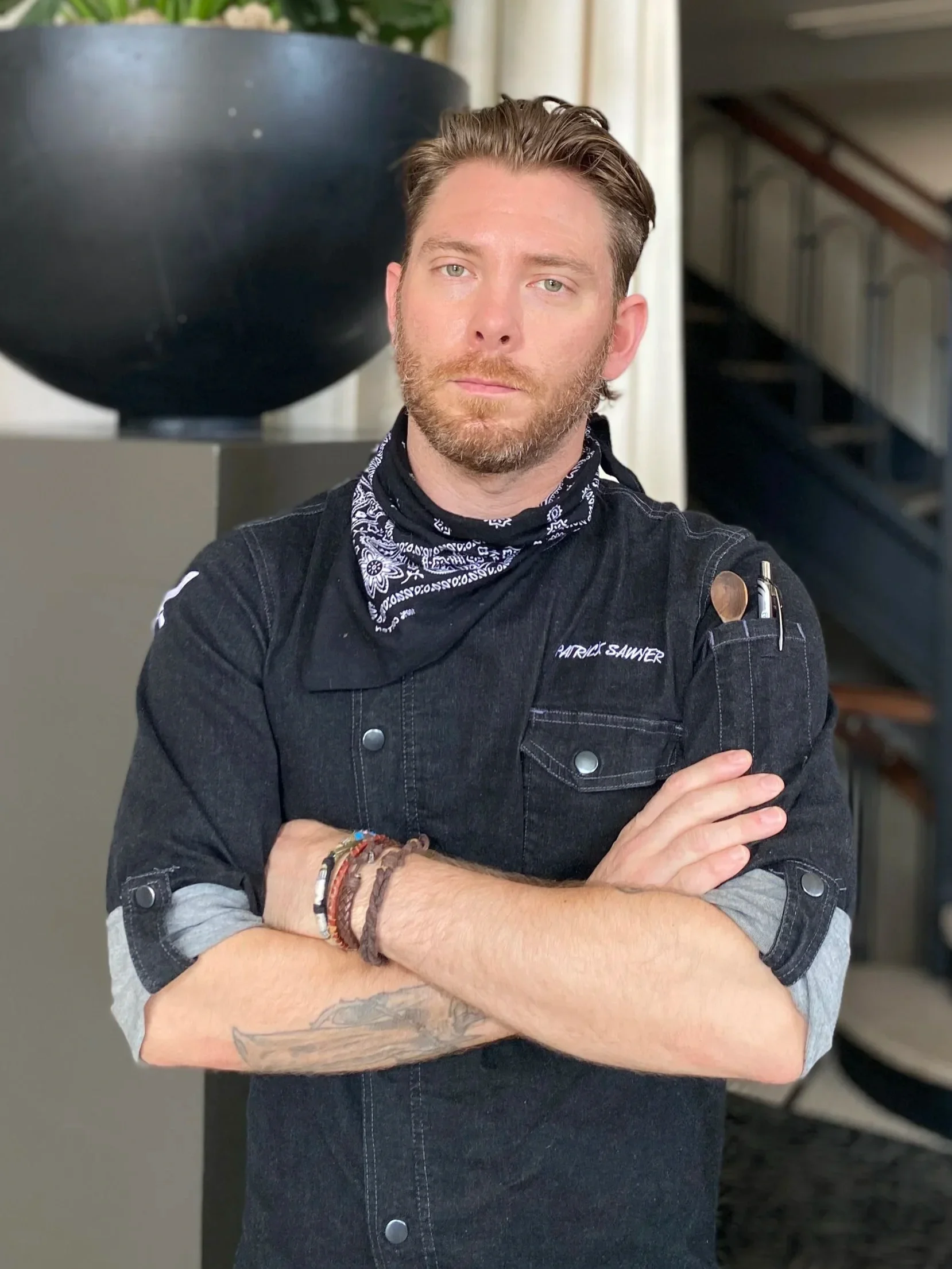 A man with brown hair and a beard standing with arms crossed in front of a large potted plant, wearing a black shirt with rolled-up sleeves, a bandana, and bracelets, indoors near stairs and curtains.