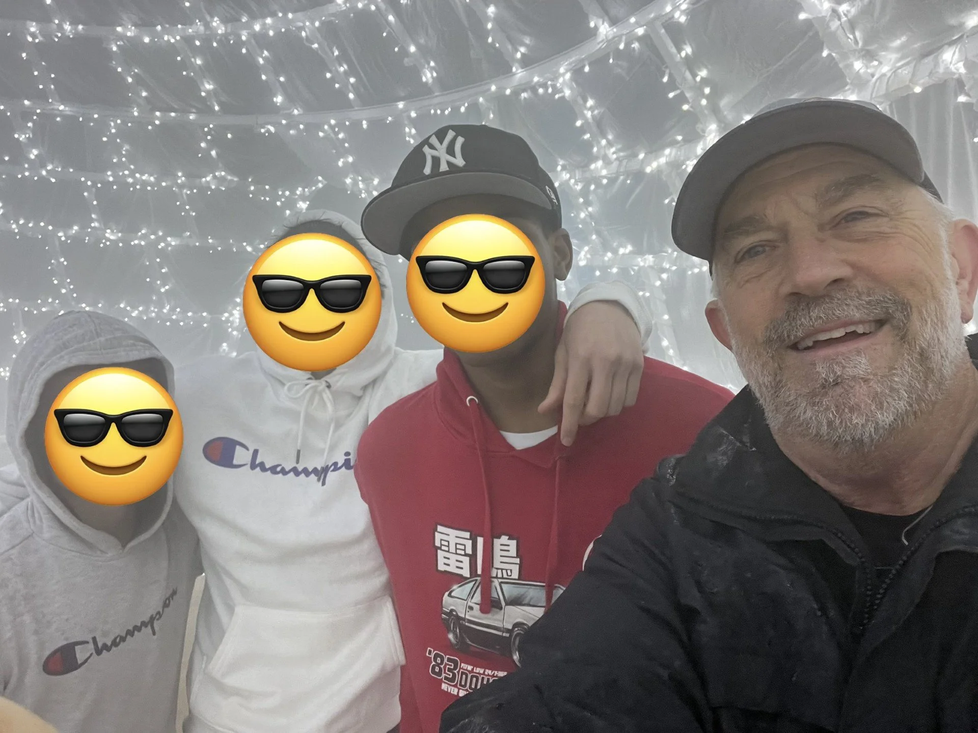 Four people taking a selfie in front of a decorated background with string lights, wearing casual clothing and sunglasses emojis covering faces for privacy.