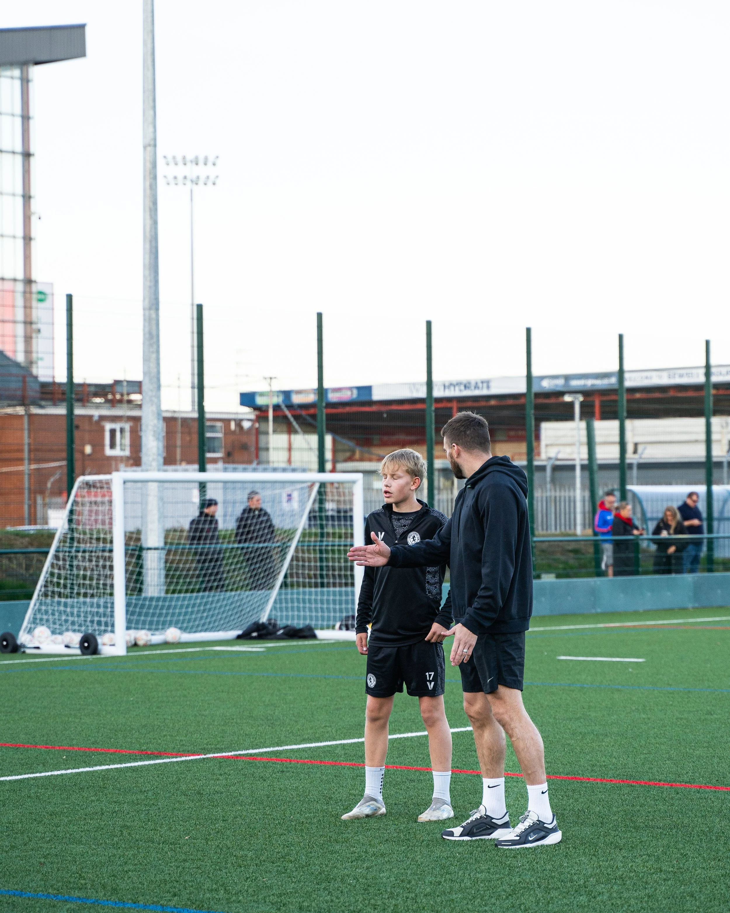 A coach and a young soccer player having a conversation on a soccer field during practice or a game, with a goal and some people in the background.