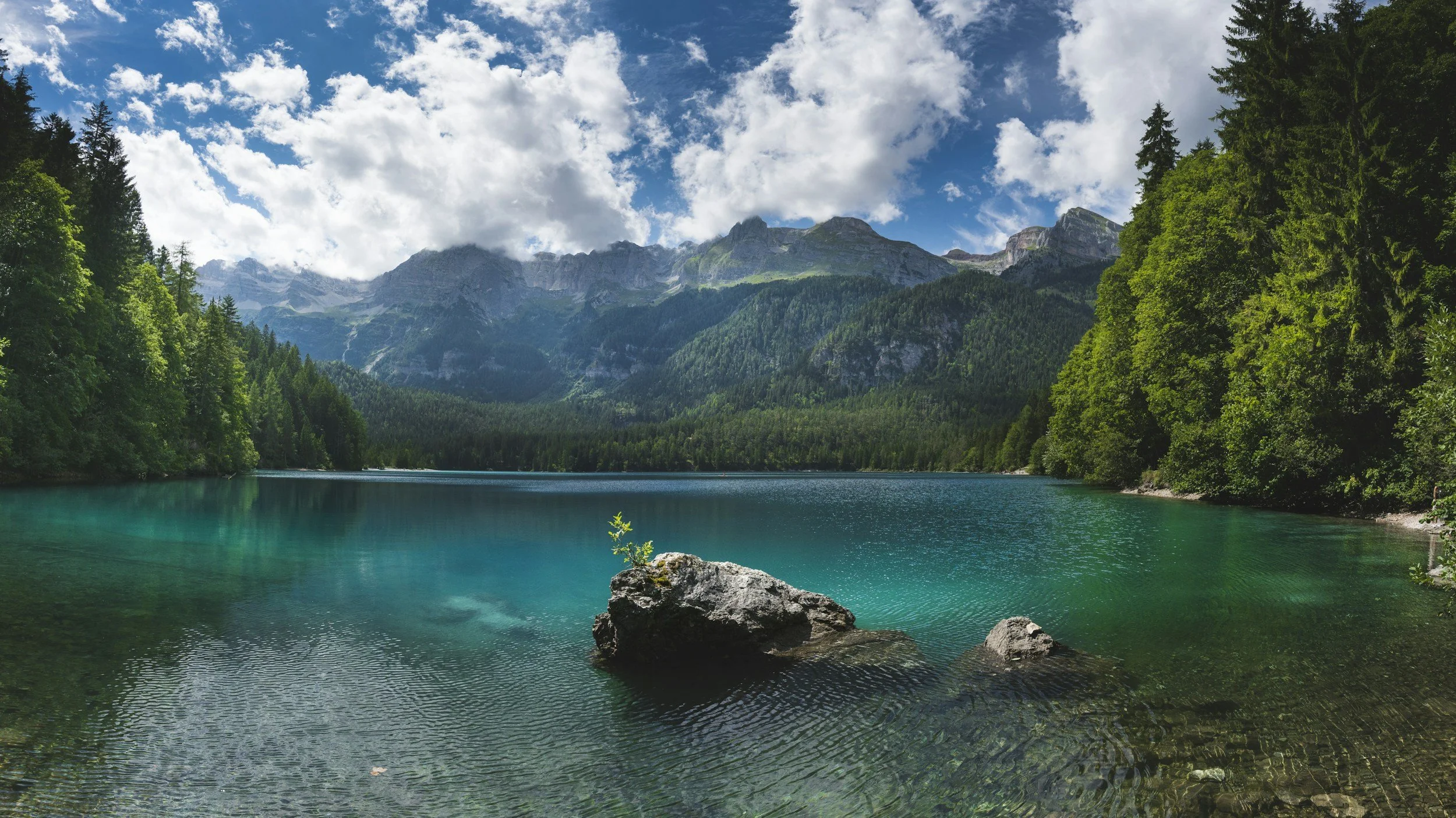 A scenic landscape of a clear turquoise lake surrounded by dense green trees and mountains under a partly cloudy sky.