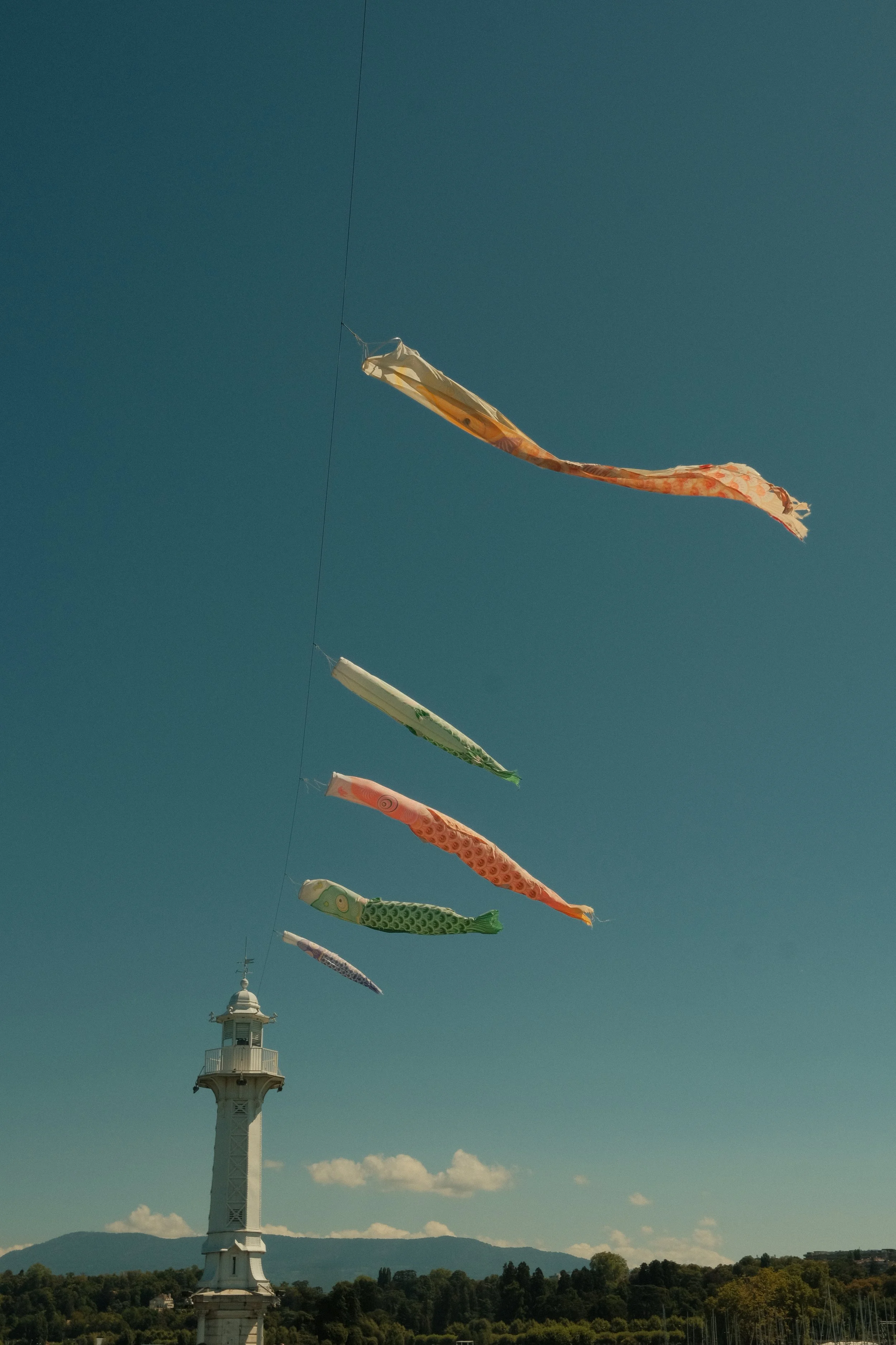 Cerf-volant en forme de poissons colorés volant dans le ciel au-dessus d'une tour blanche surplombant une zone boisée, avec des montagnes en arrière-plan.