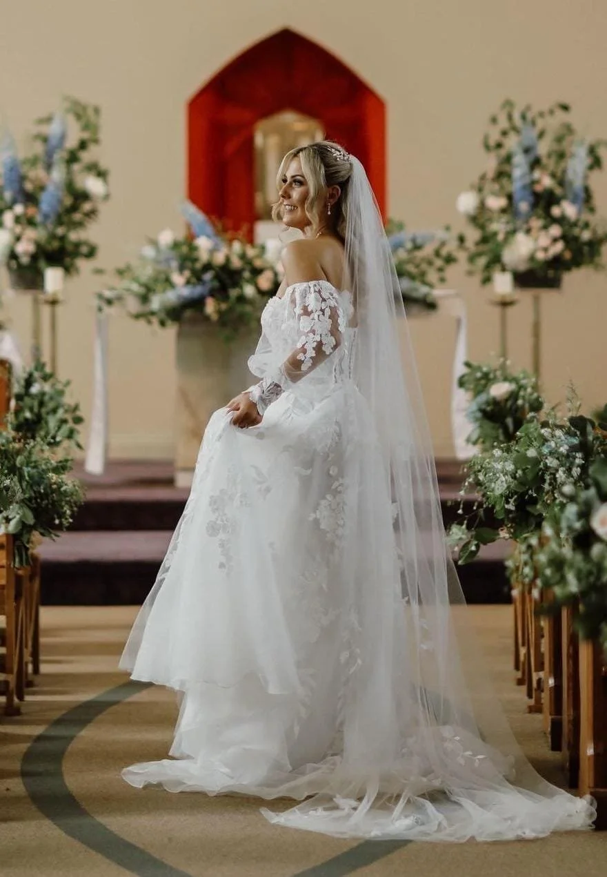 A bride wearing a lace white bridal gown, veil in her hair in a church with colorful flowers in the background 