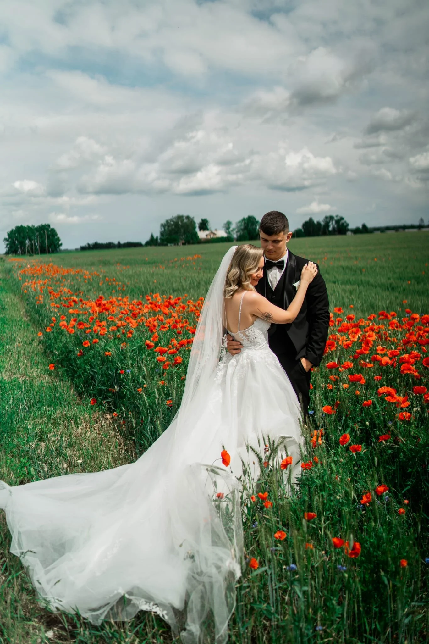 A bride wearing white gown with her groom in black tuxedo in a field of red flowers and cloudy sky 