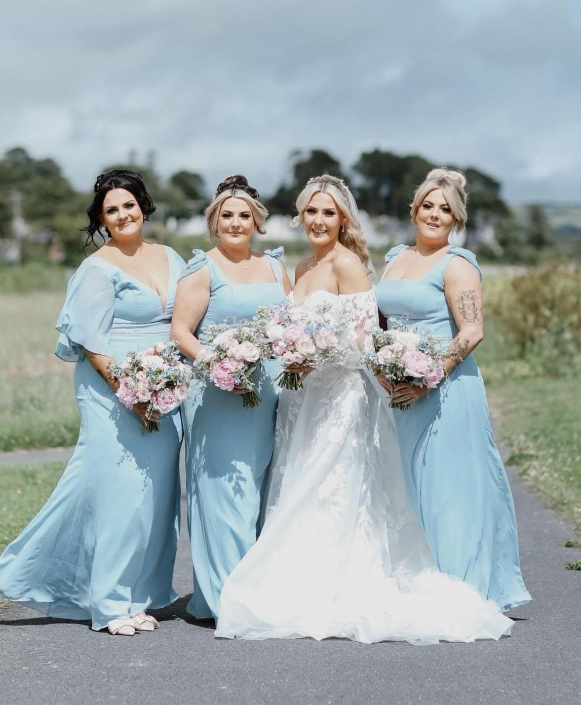 A bride in her white lace sweetheart neckline gown with her bridesmaids who are wearing blue True bridesmaids dresses and holding their pastel coloured bouquets 