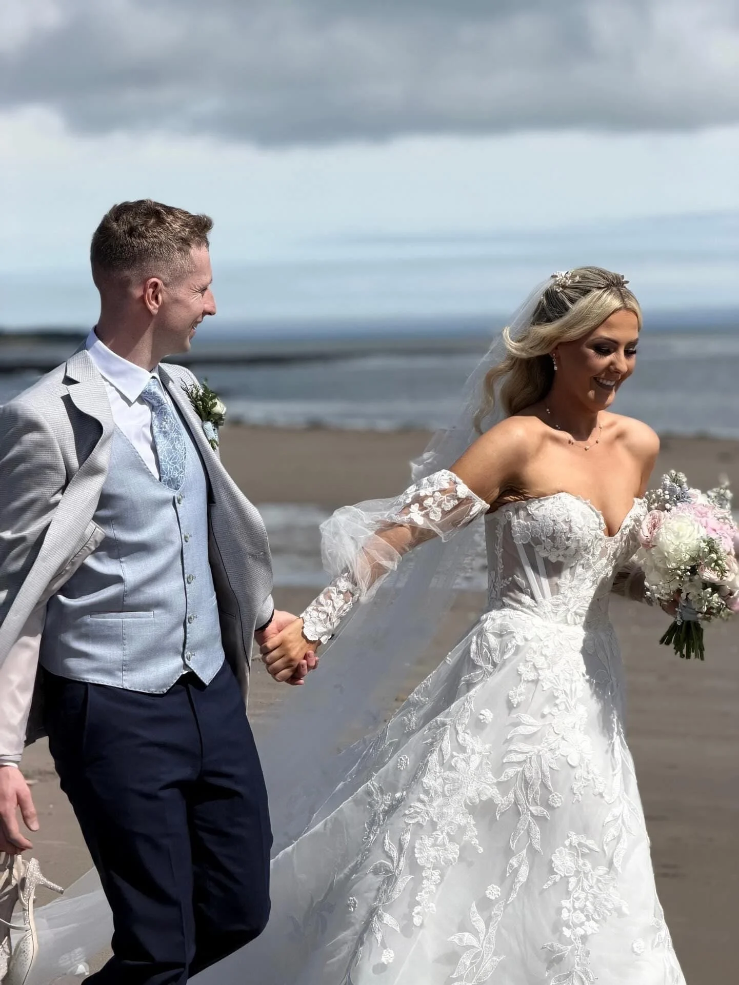 A bride in her white lace dress, sweetheart neckline and lace sleeves with her groom wearing grey and blue suit on the beach 