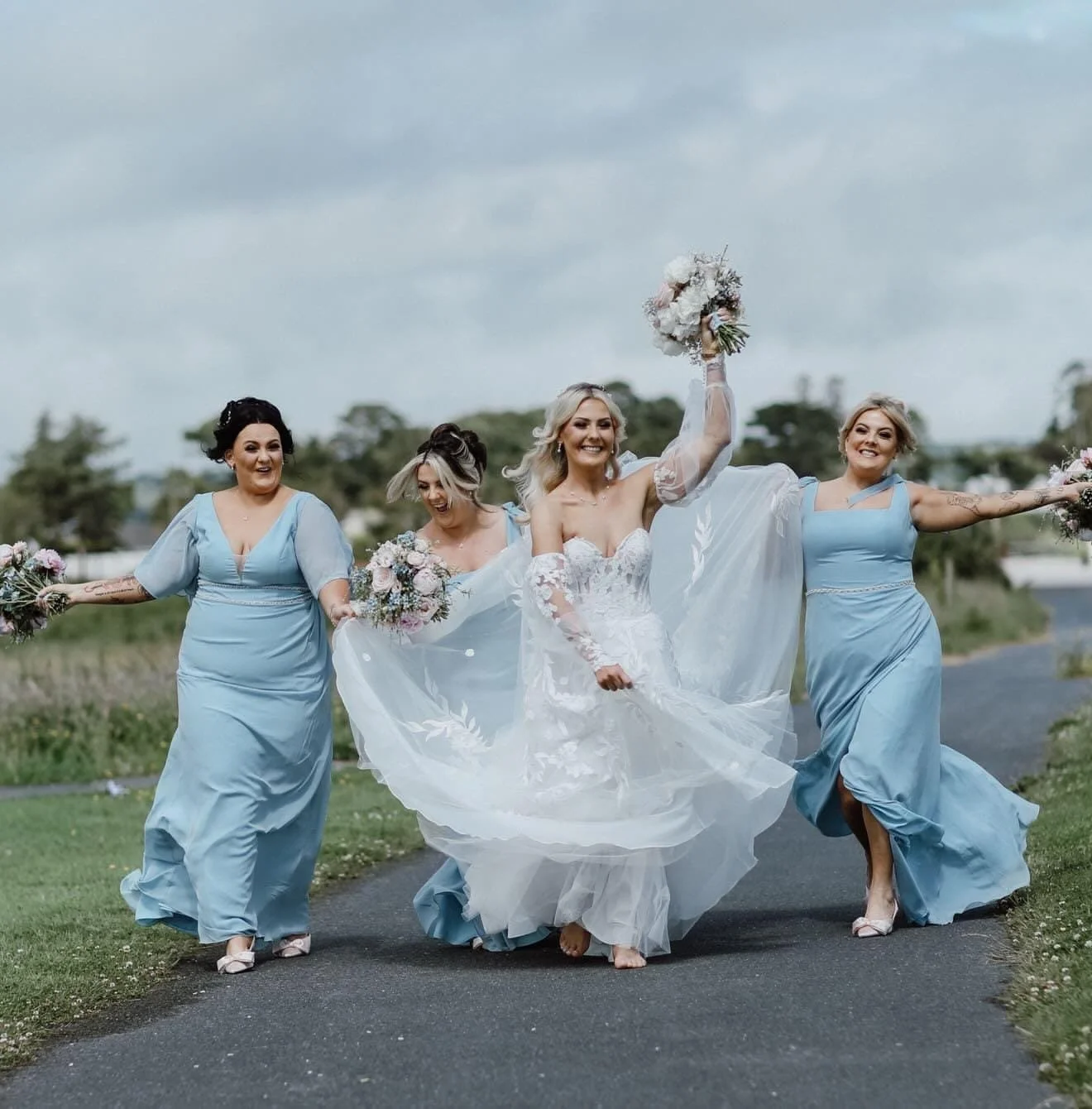 A bride in her white gown holding her bouquet in the air having fun with her bridesmaids who are wearing blue bridesmaid dresses, having fun and smiling