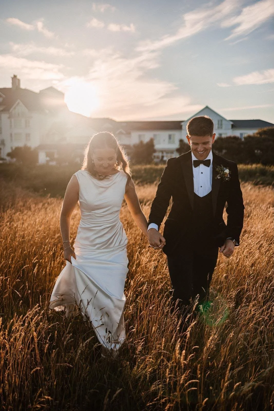 A bride in her white gown and her groom in his black tuxedo walking through a field with garryvoe hotel in the background