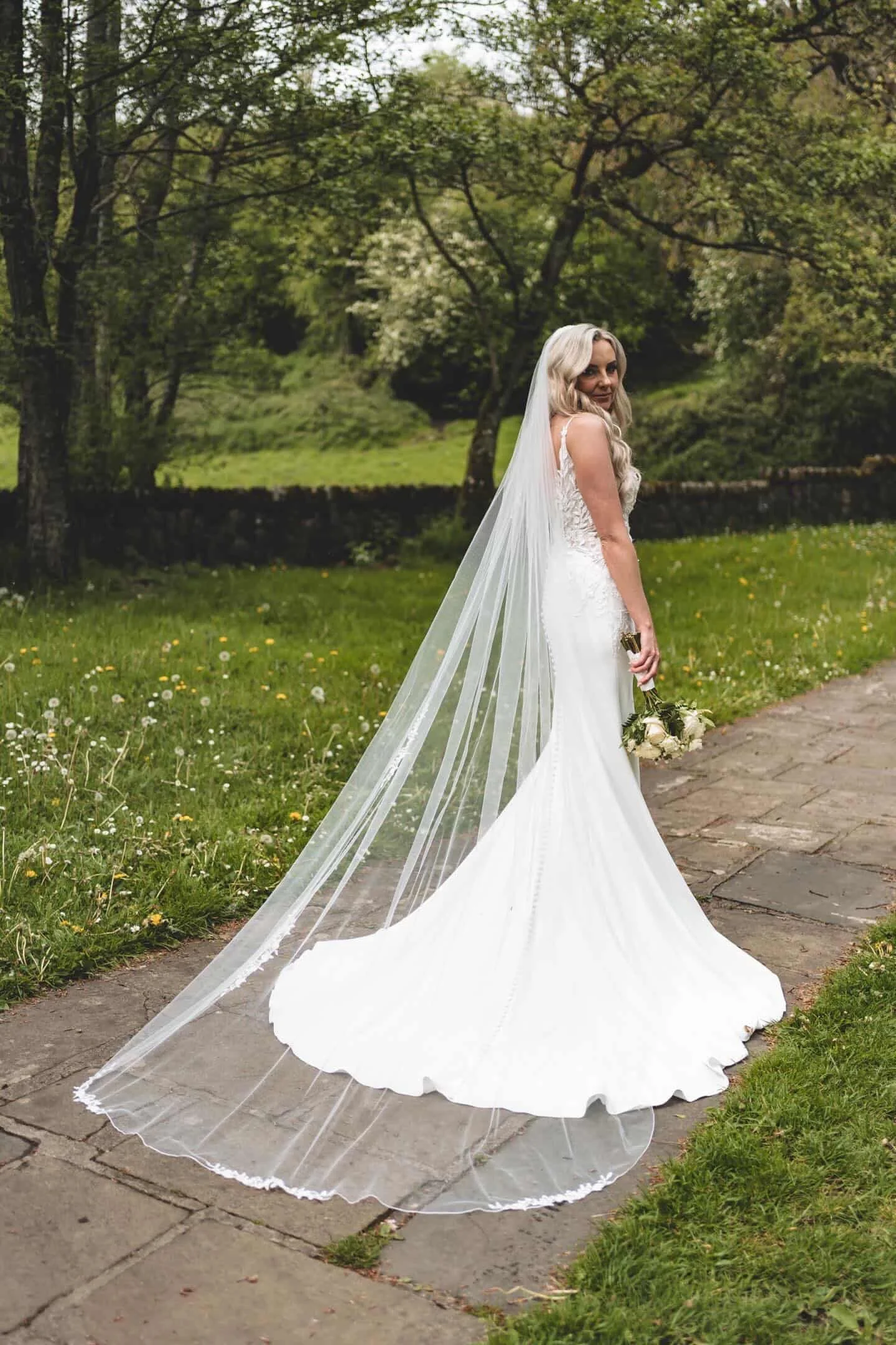 A bride in a beautiful fitted dress with a veil and train. Holding her bouquet of flowers looking over her shoulder for the photographer. Location is St. Patricks well, Clonmel, Co. Tipperary.
