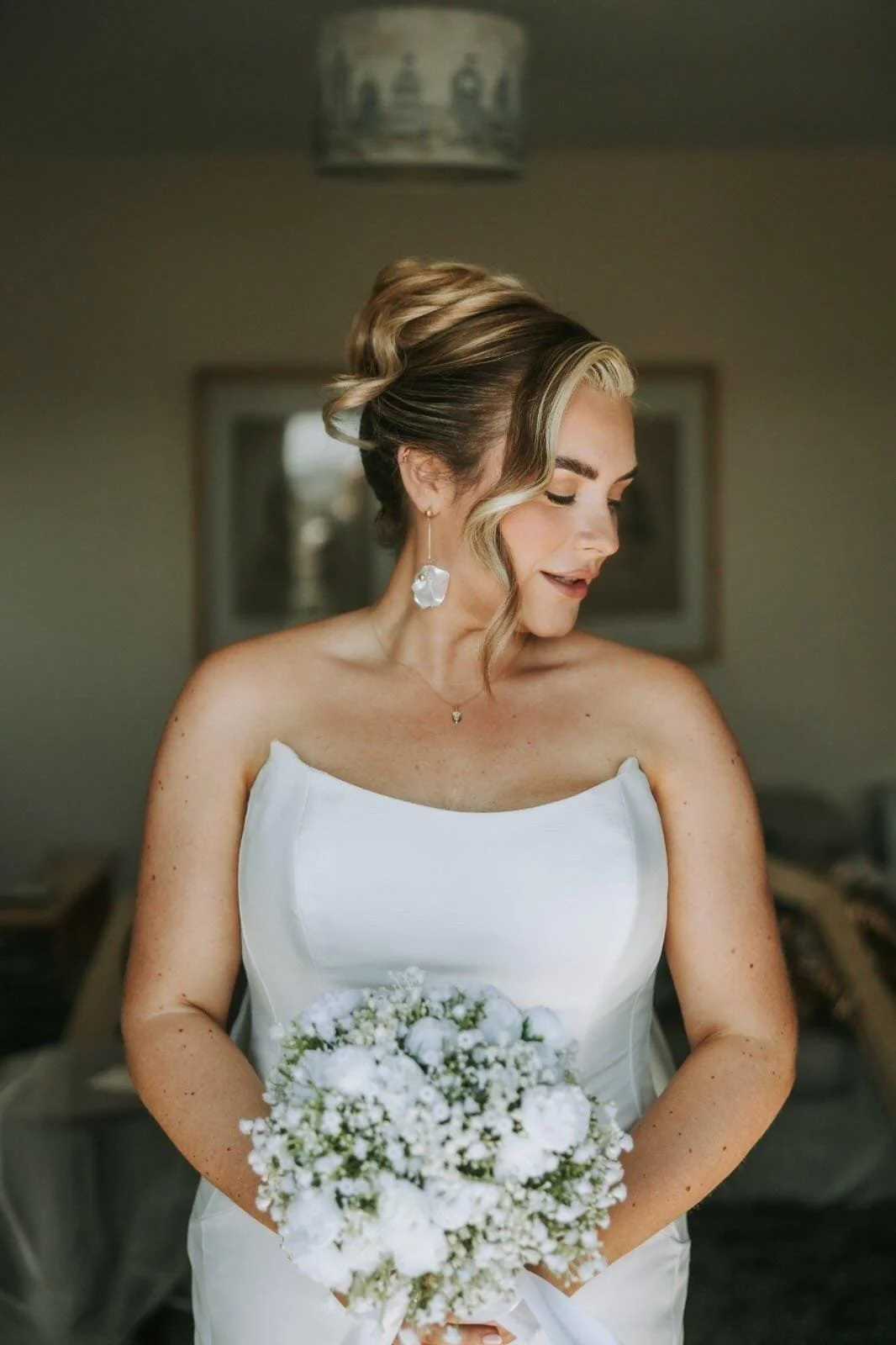 A bride in her strapless white bridal gown with her curly blonde hair and her drop earrings holding her white rose and baby's breath bouquet