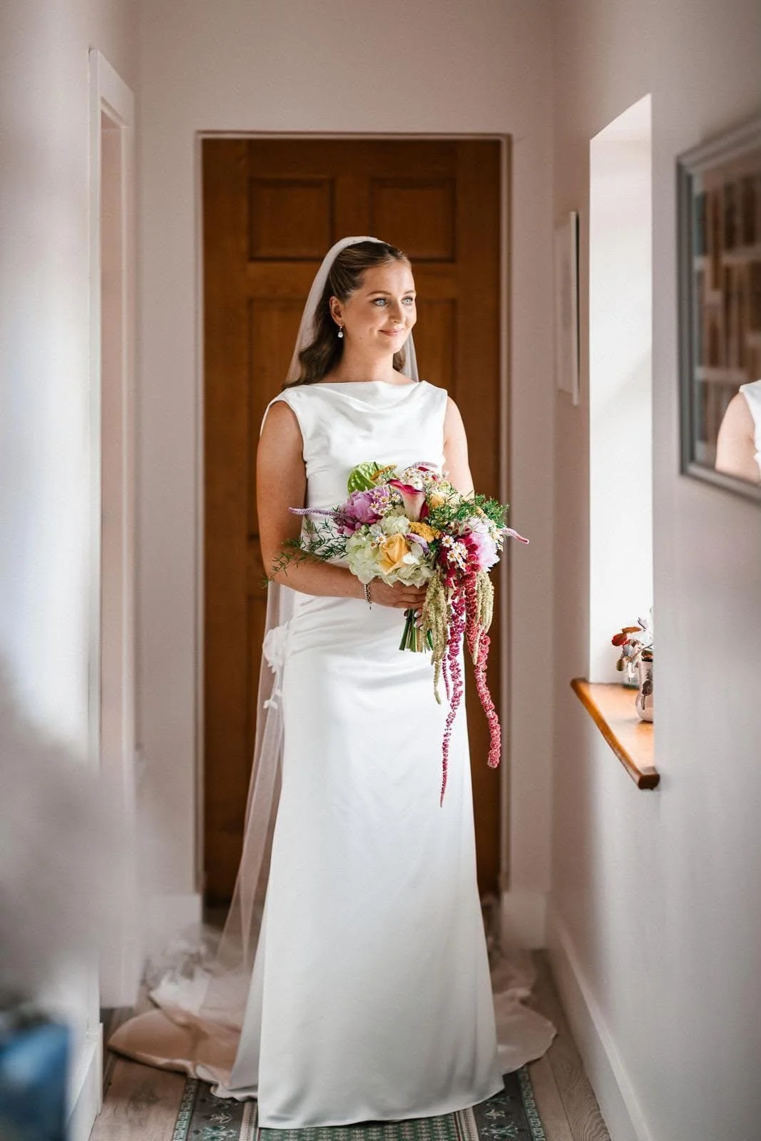 A Bride in white dress holding colourful bouquet wearing a veil in hallway looking out window