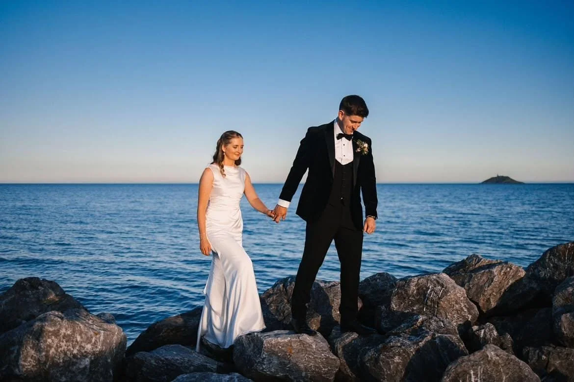 A bride in white gown and groom in black tuxedo walking on rocks blue sky and sea in background