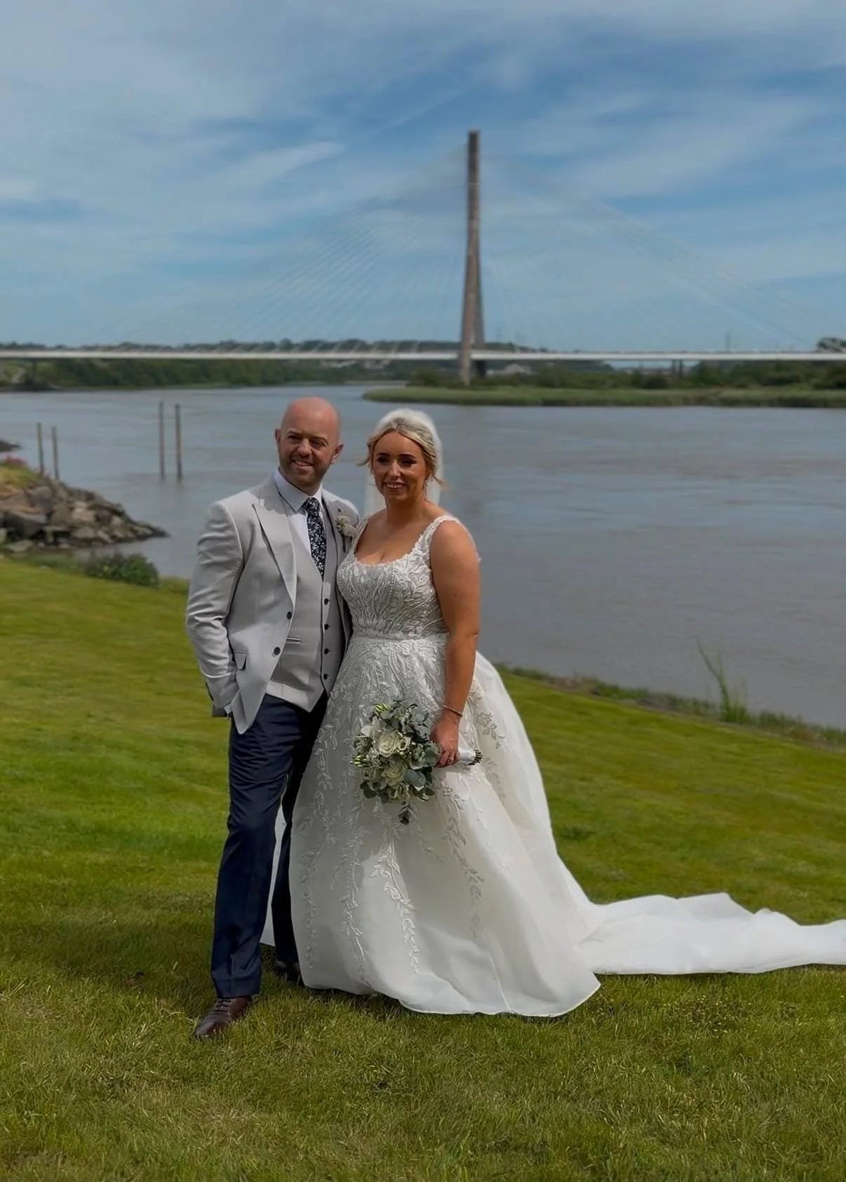 A Bride in white gown holding colourful bouquet with her groom in grey suit and navy trousers and waterford bridge in background