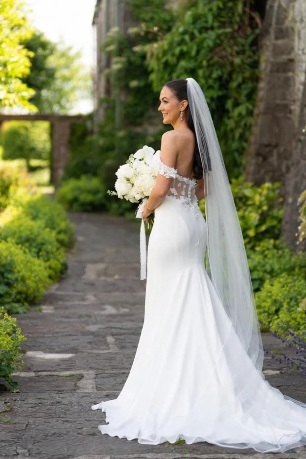 Bride in beautiful Mikaella white dress, holding white rose bouquet, veil in hair with green flowers in background