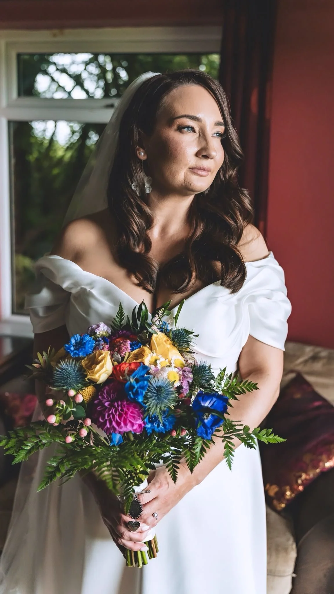 A bride with brown curly hair wearing white dress holding beautiful colourful bouquet looking out window