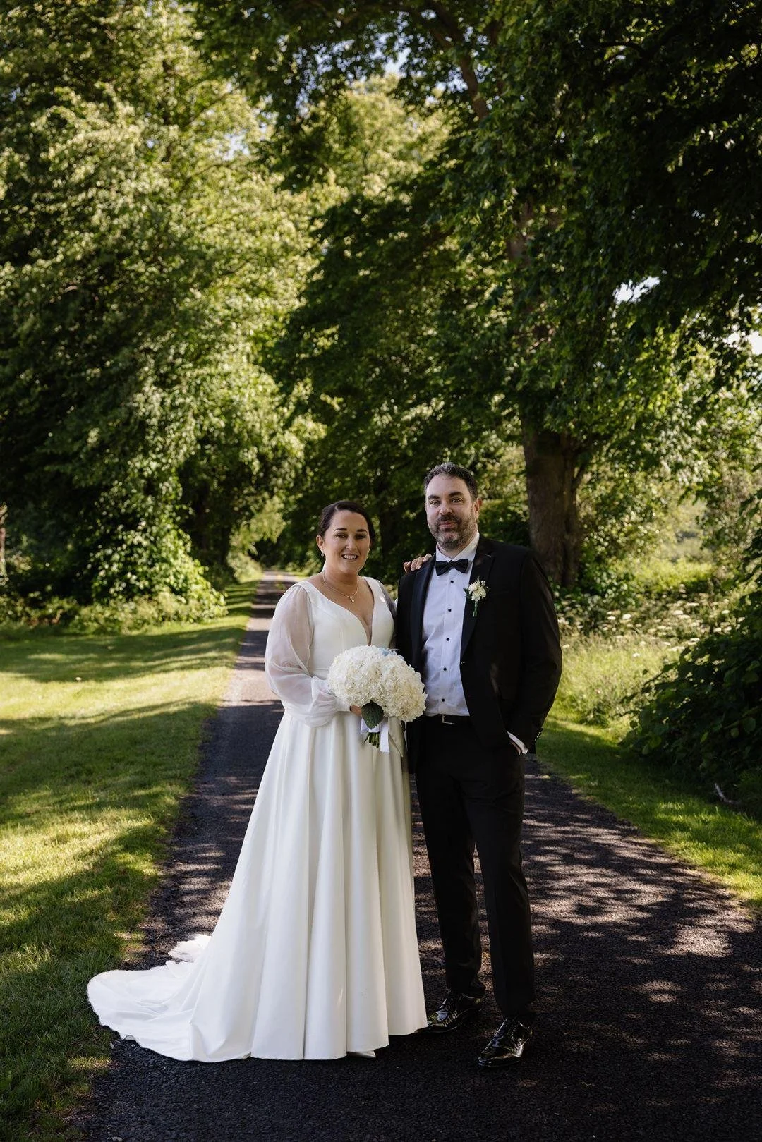 A Bride wearing a white gown and her groom wearing a black tuxedo holding her white bouquet with trees in background