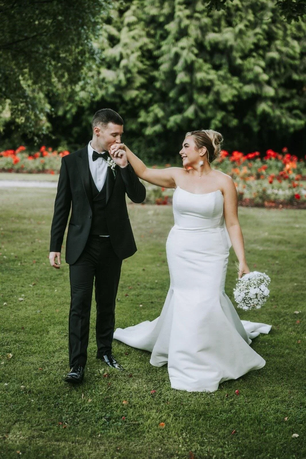 A Bride in white dress and her groom in black tuxedo kissing her hand on the grounds of faithlegg house in waterford with her white bridal bouquet