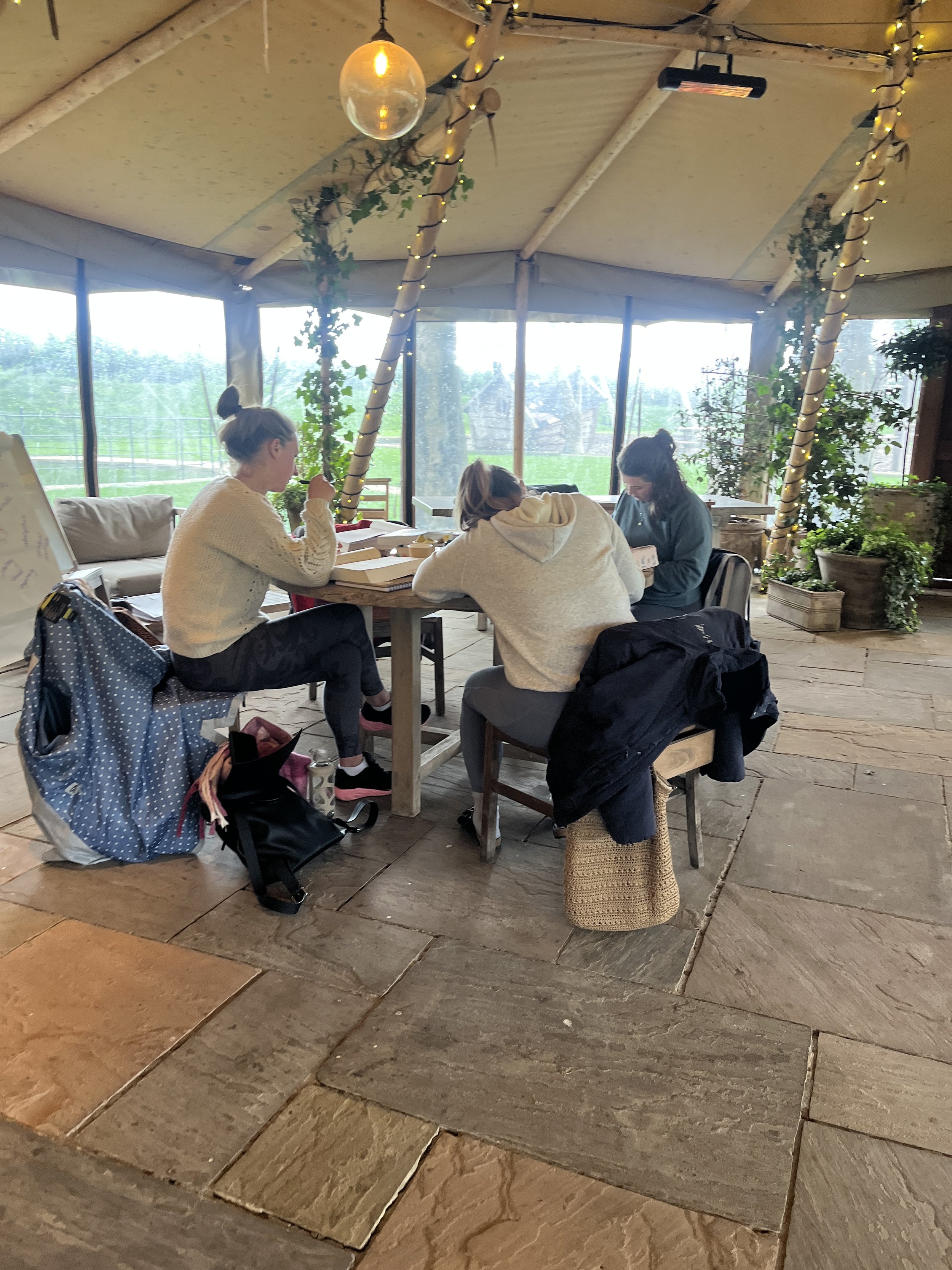 Three young women sitting at a rustic indoor table, reading or working. The room has large windows overlooking a green outdoor area. Decor includes string lights and leafy plants. The women are dressed casually, and there are backpacks and bags at their feet.