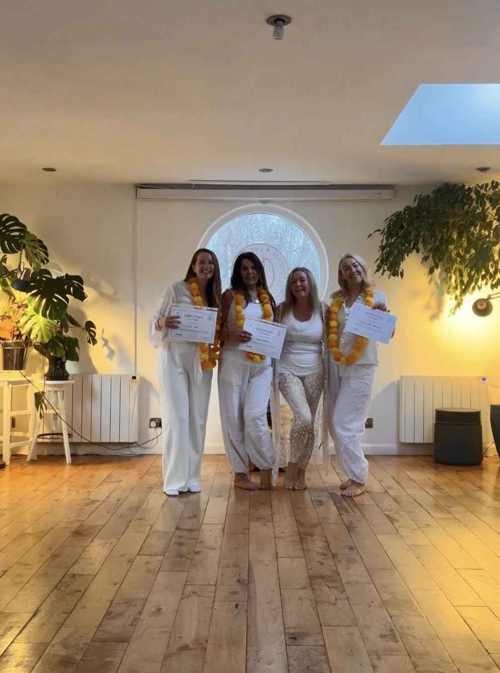 Four women in white clothing and yellow-orange leis standing in a room with wooden floors, holding certificates, smiling, with large plants and a circular window behind them.
