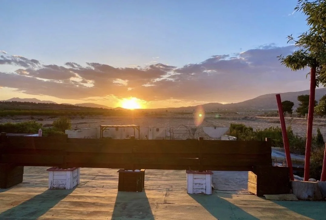 Sunset over a rural landscape with mountains in the distance, trees, and a clear sky with some clouds. A wooden fence, two white painted crates, and some red poles are in the foreground, with a wooden deck or platform.