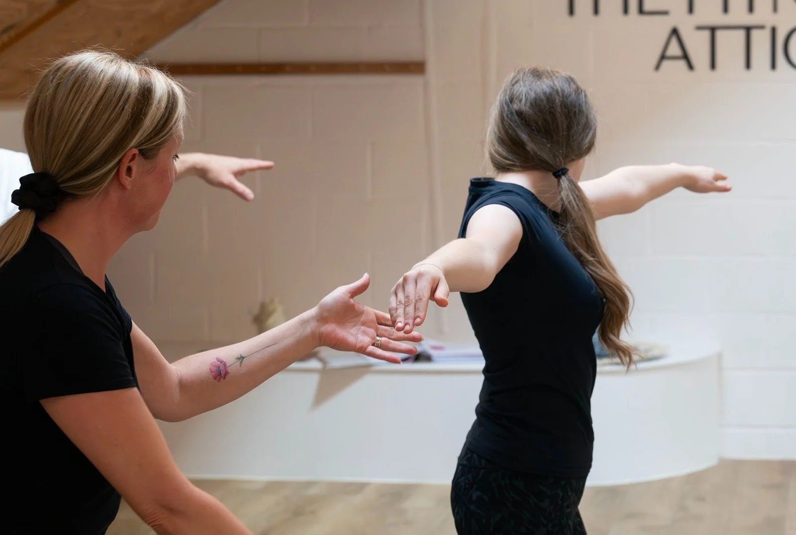 A woman with blonde hair tied in a ponytail assisting a young girl in practicing a yoga pose in a studio. The girl is extending her arms out to the sides and slightly forward while the woman is guiding her. There is a part of a wall with written text visible in the background.