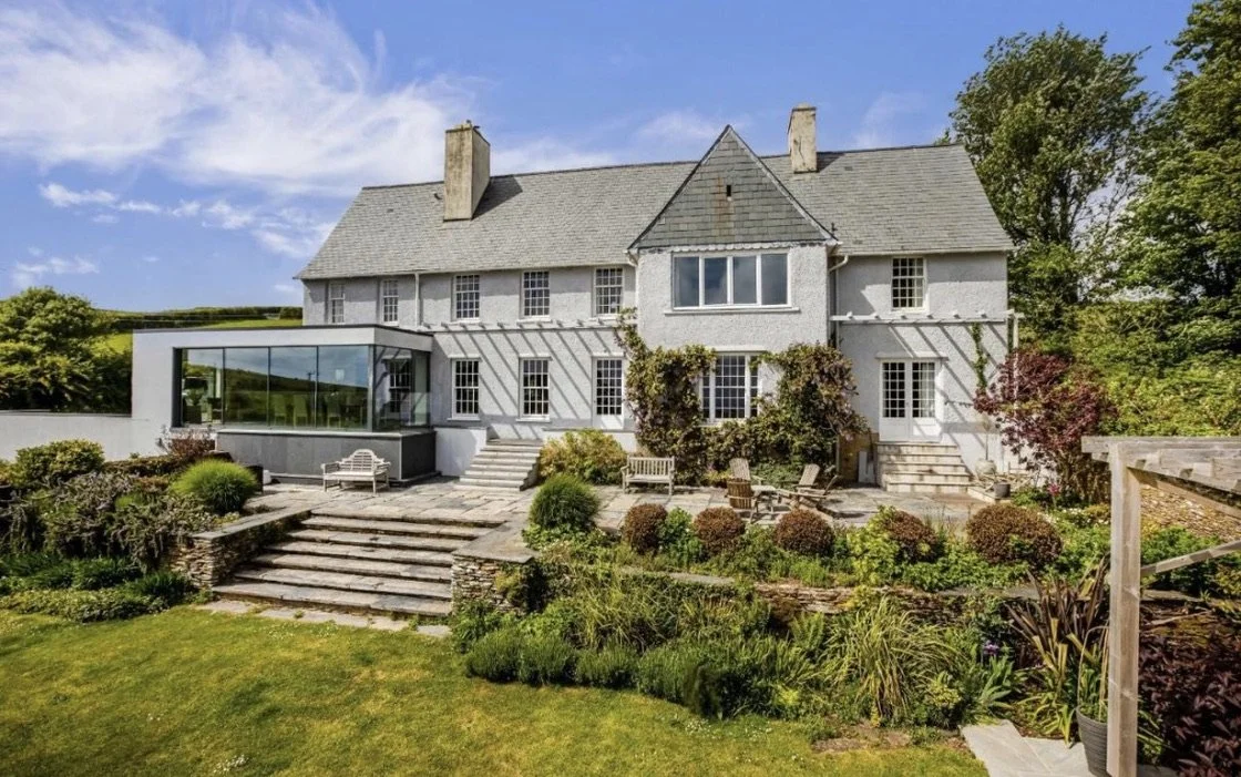 Large white house with gray roof and multiple chimneys, surrounded by lush greenery and landscaped garden with stone stairs and outdoor seating.