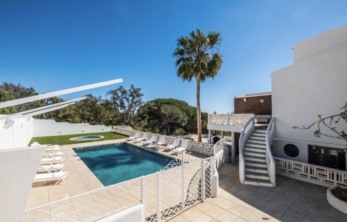 A luxurious backyard with a swimming pool, poolside lounge chairs, a grassy area with a small circular hot tub, a tall palm tree, and a clear blue sky.