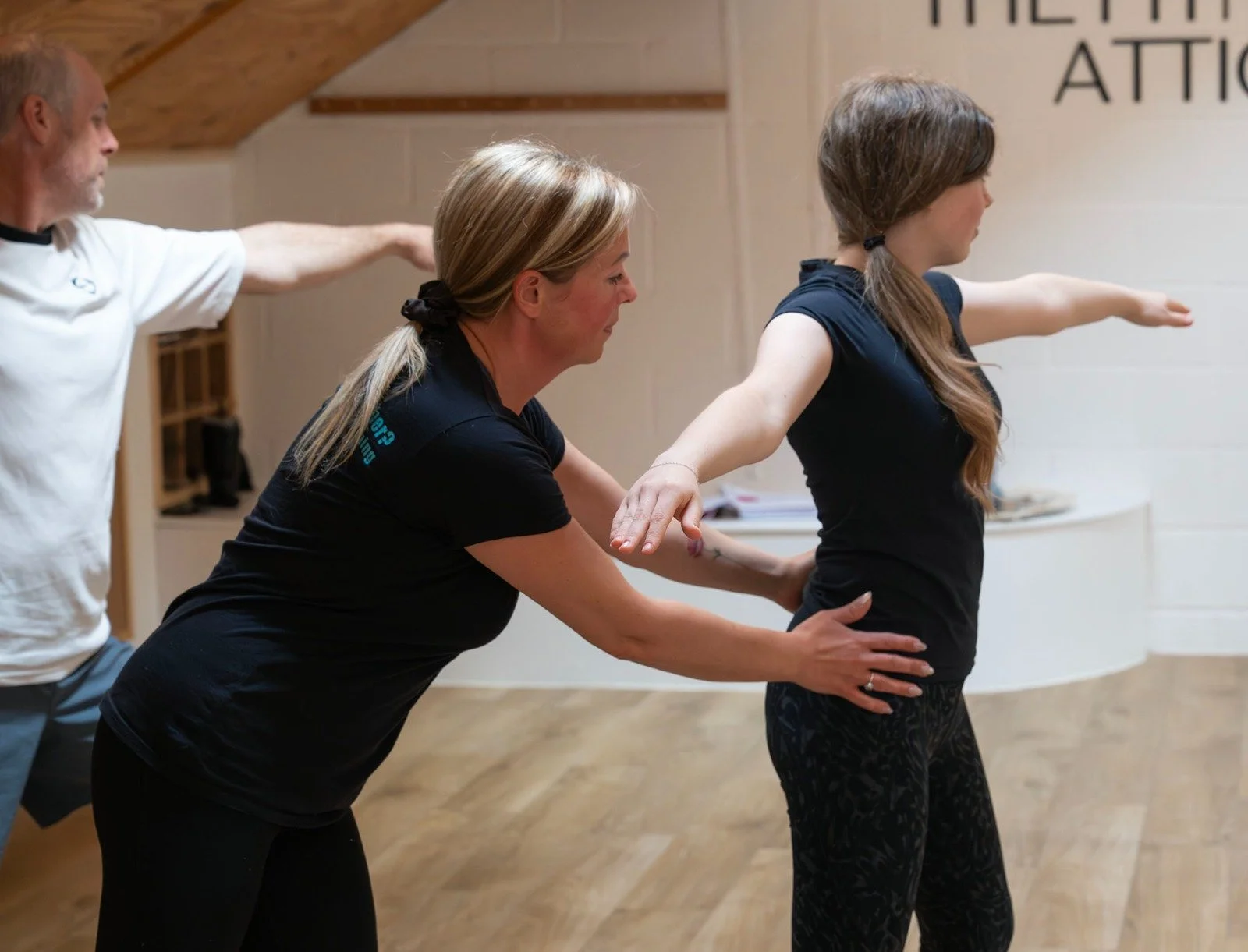 Three people participating in a physical therapy or exercise class, with two women assisting a young woman with stretching arm movements in a room with wooden flooring and a wall with writing.