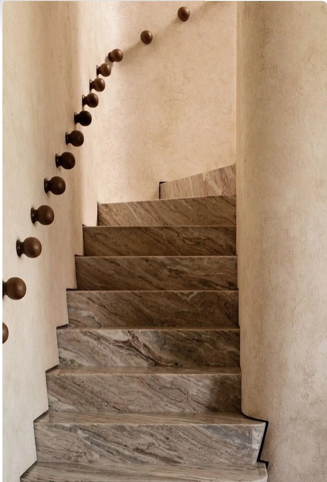 Marble staircase with a curved wall on the right and a beige textured wall on the left, featuring a vertical row of small, round wooden knobs.