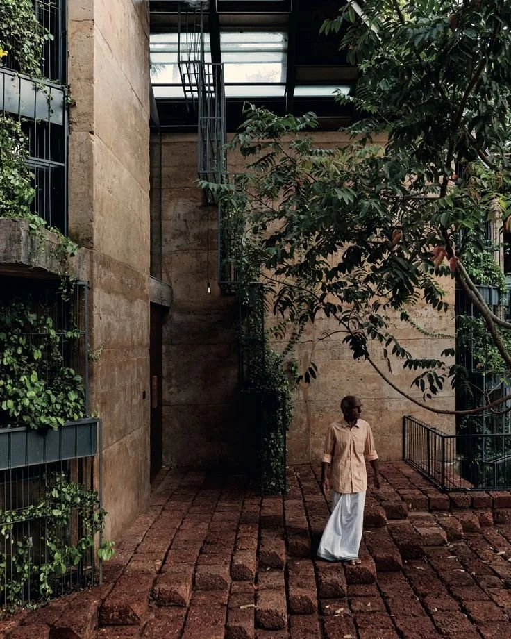 A man standing on a brick pathway outdoors next to a concrete building, with green trees and plants around, under a partially open metal roof.