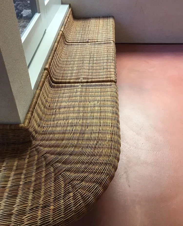 A wicker bench with three cushions near a window in a room with pinkish floors and beige walls.