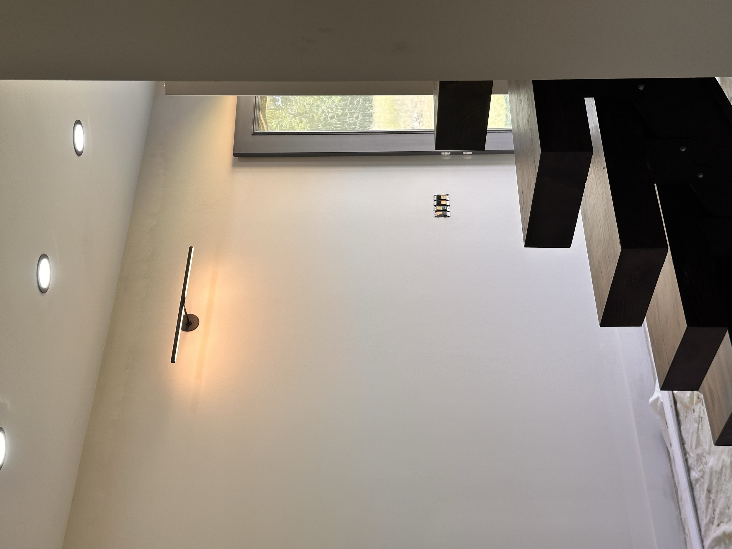 Interior of a modern room with white walls, a large window with a view of greenery, six recessed ceiling lights, and a black wooden staircase.