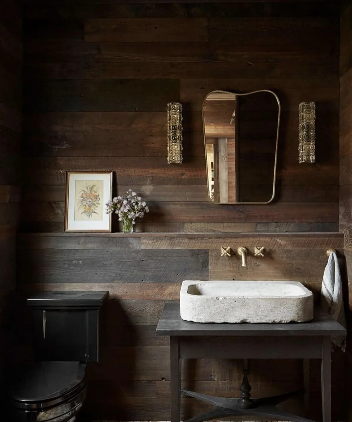 A rustic bathroom featuring dark wooden wall paneling, a cream-colored stone vessel sink on a wooden vanity, gold fixtures, an oval mirror, two decorative wall sconces, a framed floral artwork, a vase with flowers, a black toilet, and a towel hanging