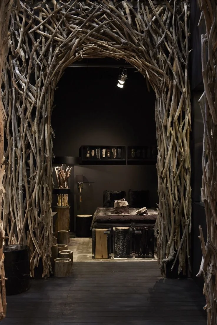 Interior of a room seen through a rustic archway made of intertwined branches. The room features a bed with dark bedding, side tables, and wall-mounted shelves with decorative items, all in a dark-themed decor.
